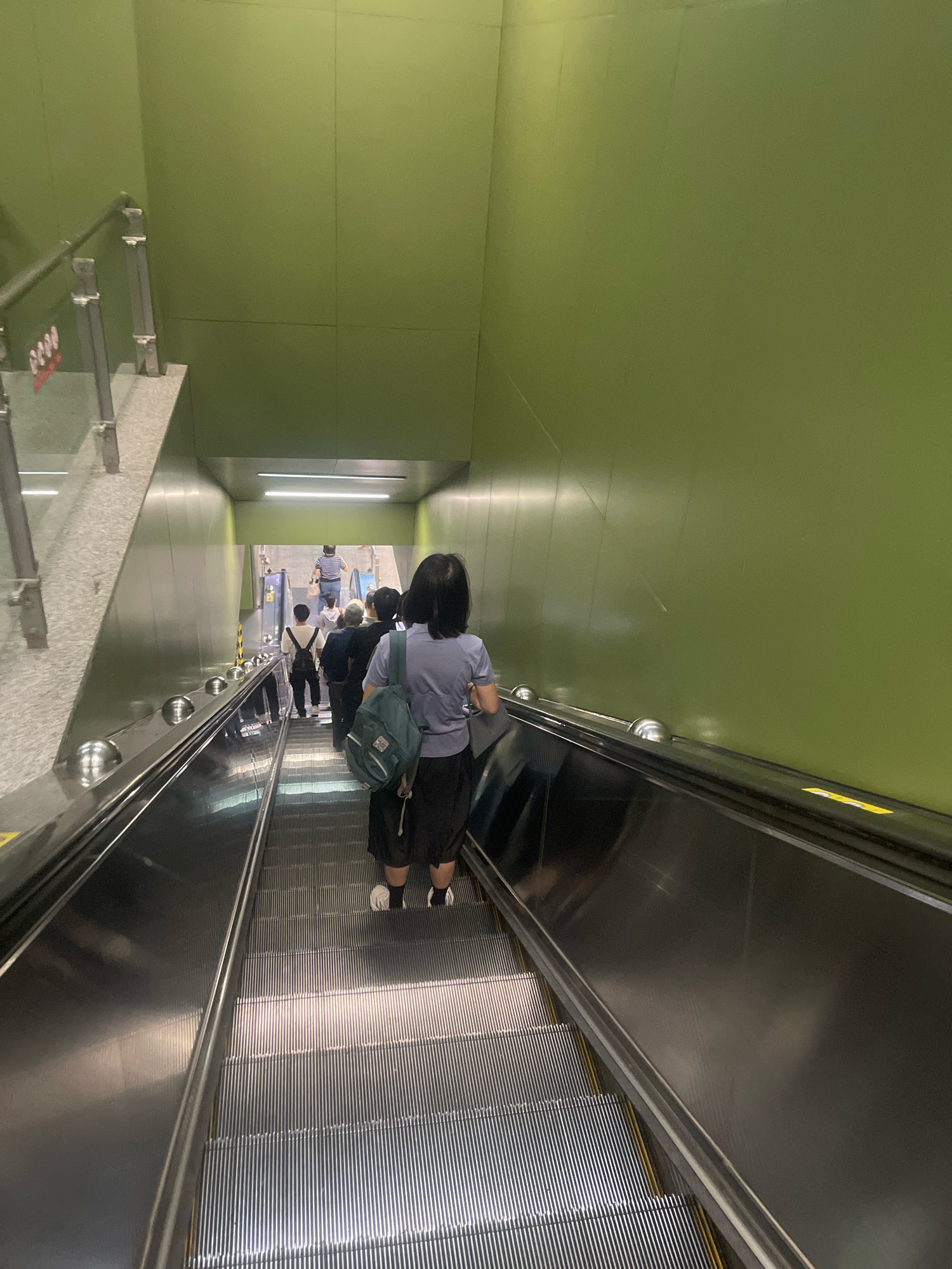 The image shows a view from an escalator in a public space, likely a subway station in Yuexiu District, China. The walls of the escalator tunnel are painted a bright green. Several people are visible on the escalator, all moving downwards. In the foreground, a woman with a backpack is facing away from the camera, and to her left, the escalator steps are clearly visible. Further down the escalator, a line of people is waiting or moving, and at the bottom, more individuals are seen on a flat surface. The lighting is bright, with some reflections on the escalator's metal surfaces.