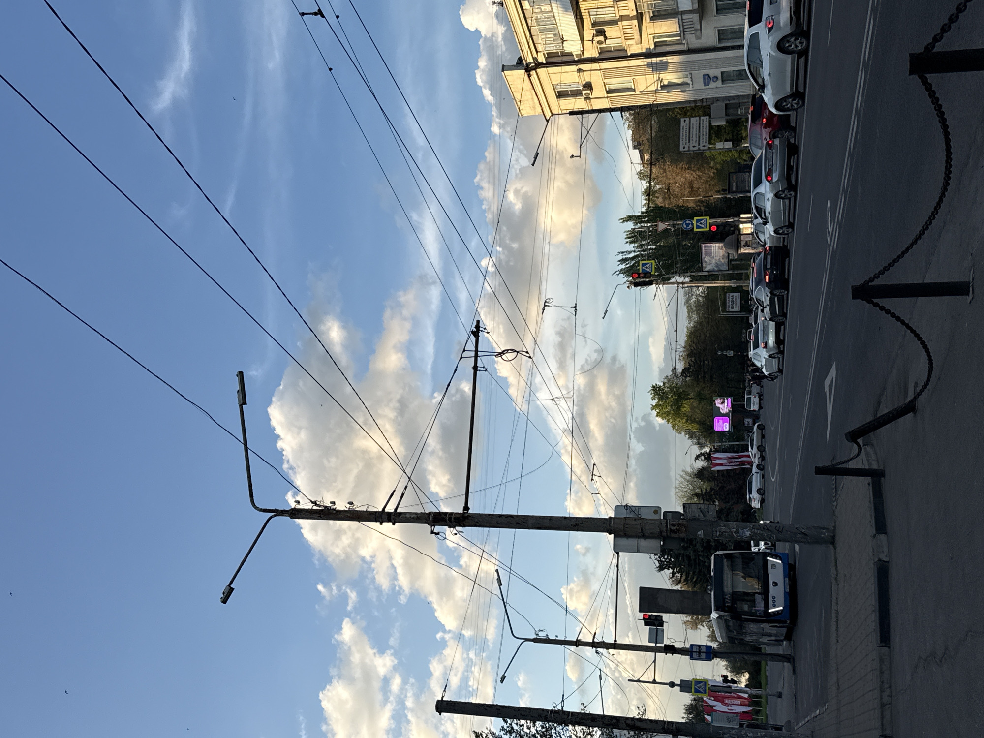 The image shows a street scene in Rostov-on-Don, Russia. The view is angled upwards, showcasing a cloudy sky intersected by a network of overhead power lines and utility poles. A multi-story building stands along the right side of the frame with vehicles parked along the roadside. A line of vehicles is visible, indicating traffic congestion. In the foreground, a bus and a sidewalk with a chain fence can be seen. Traffic lights are red.