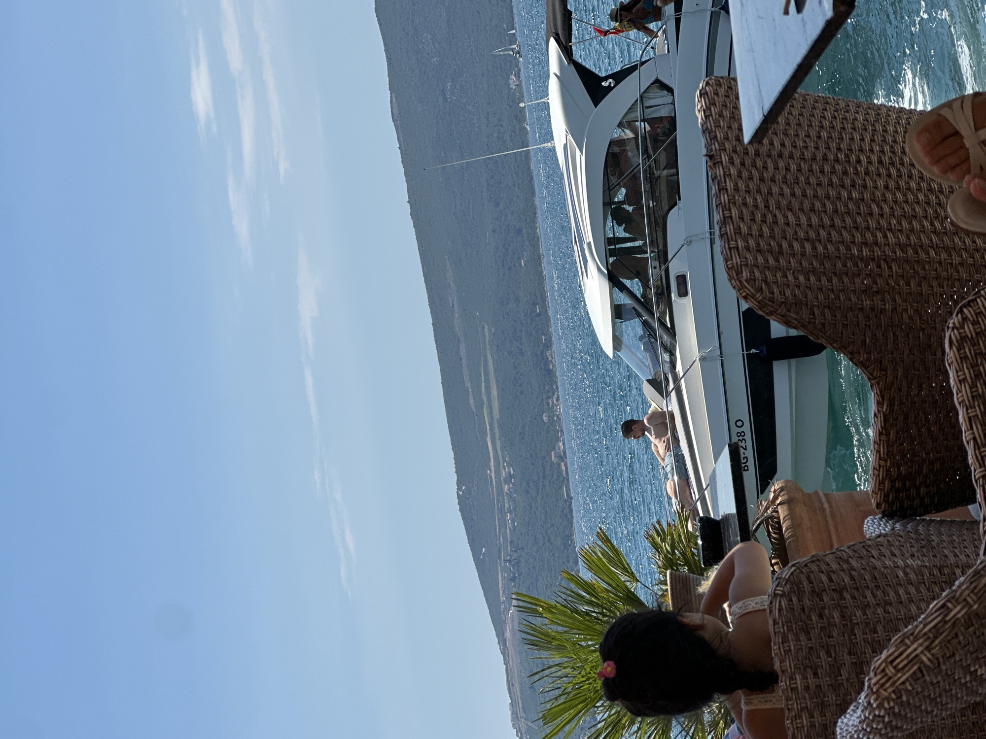 In Donja Lastva, Montenegro, on a sunny day, a white motorboat with several passengers glides across the blue water. A man is lounging on the deck of the boat. In the foreground, two wicker chairs face the water, and a woman with her back to the camera is seated in one of them, looking towards the boat. A palm frond is visible to the left of the woman. In the background, a lush green, hilly coastline dotted with buildings rises from the water under a clear blue sky.