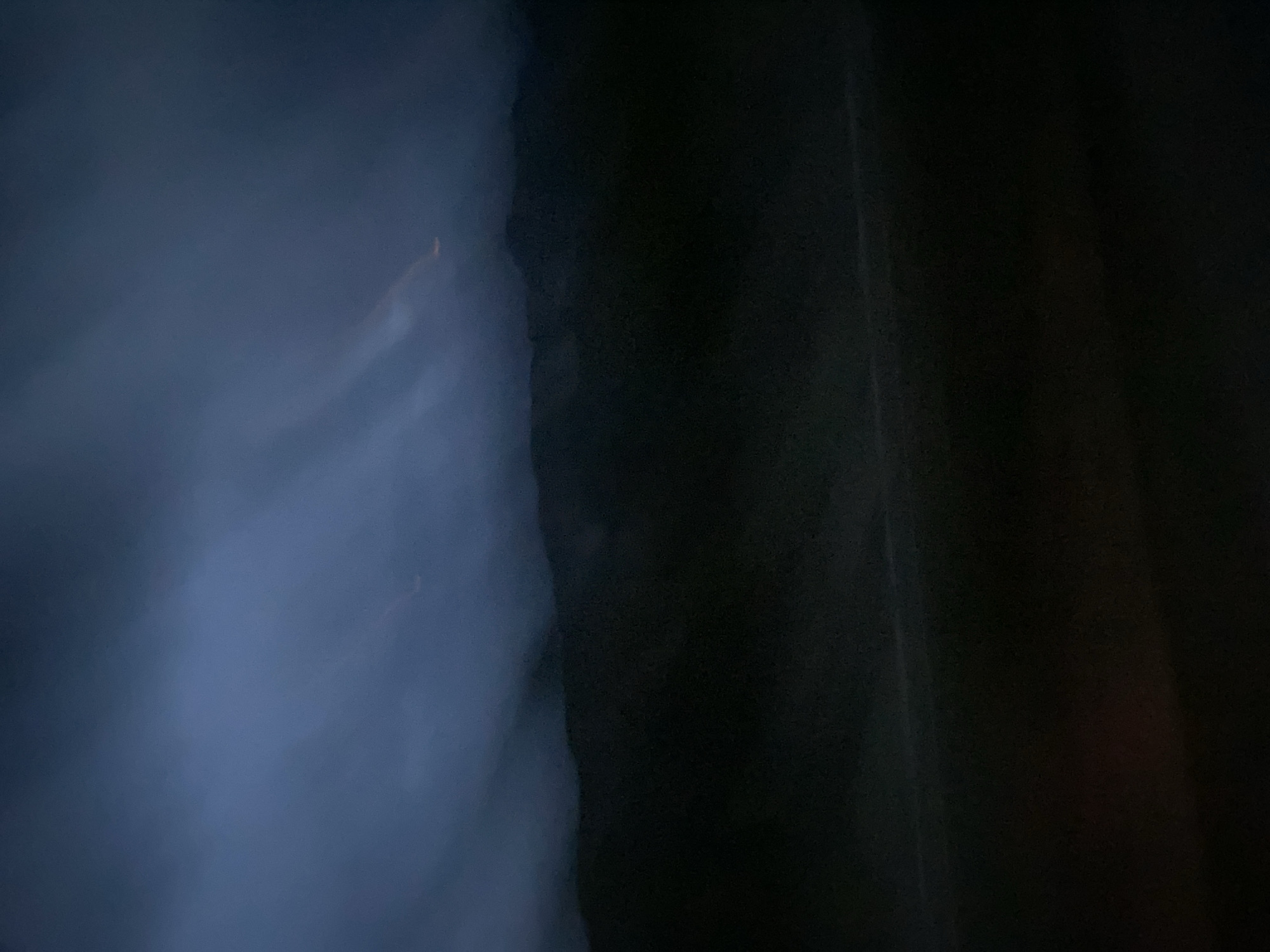 The image depicts a dark, atmospheric scene with indistinct mountain ranges silhouetted against a cloudy, twilight sky. Streaks of light, possibly from distant lightning or artificial illumination, are visible within the clouds. The foreground is shrouded in deep shadow, obscuring any details of the terrain or potential man-made structures. The overall impression is one of remoteness and natural beauty, possibly captured at dusk or during a storm. There are no discernible people or specific objects that would allow for identification of the exact location within Radfeld, Austria, beyond the general impression of a mountainous landscape. The image's low light and blurred elements contribute to its abstract and somewhat ominous quality.