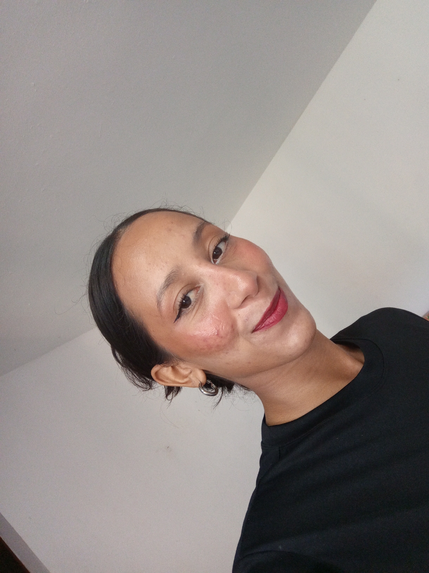 A woman with dark hair pulled back is pictured in a selfie. She is wearing dark lipstick, eyeliner, and blush. She is wearing a black shirt. The background consists of white walls. The image does not contain any information to identify the location as Porlamar, Venezuela.