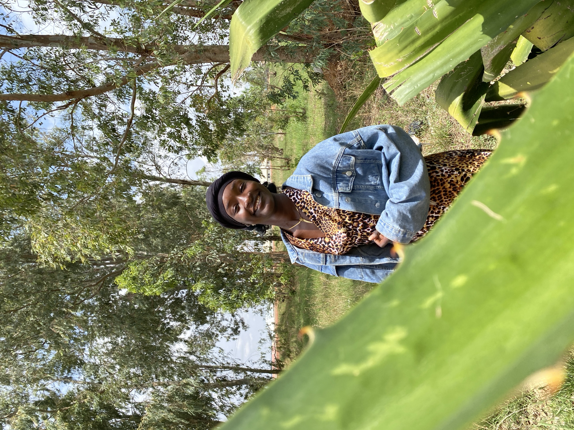 A woman wearing a black head covering, a leopard-print top, and a blue denim jacket smiles at the camera. The background includes trees and grassy ground. Large green leaves are in the foreground, partially obscuring the view. The location is outdoors, possibly a park or garden, in Durimi, Nigeria.