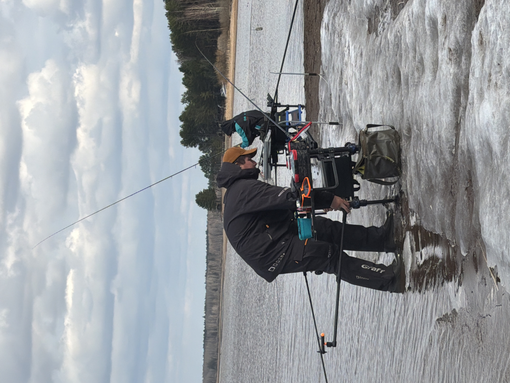 The image shows two people fishing on a lake in Chaykovsky, Russia. They are sitting on specialized fishing chairs with equipment attached, near a shoreline with ice formations. One person is wearing a brown cap and black Graff-branded clothing. The sky is cloudy. A forest is visible in the background across the water.