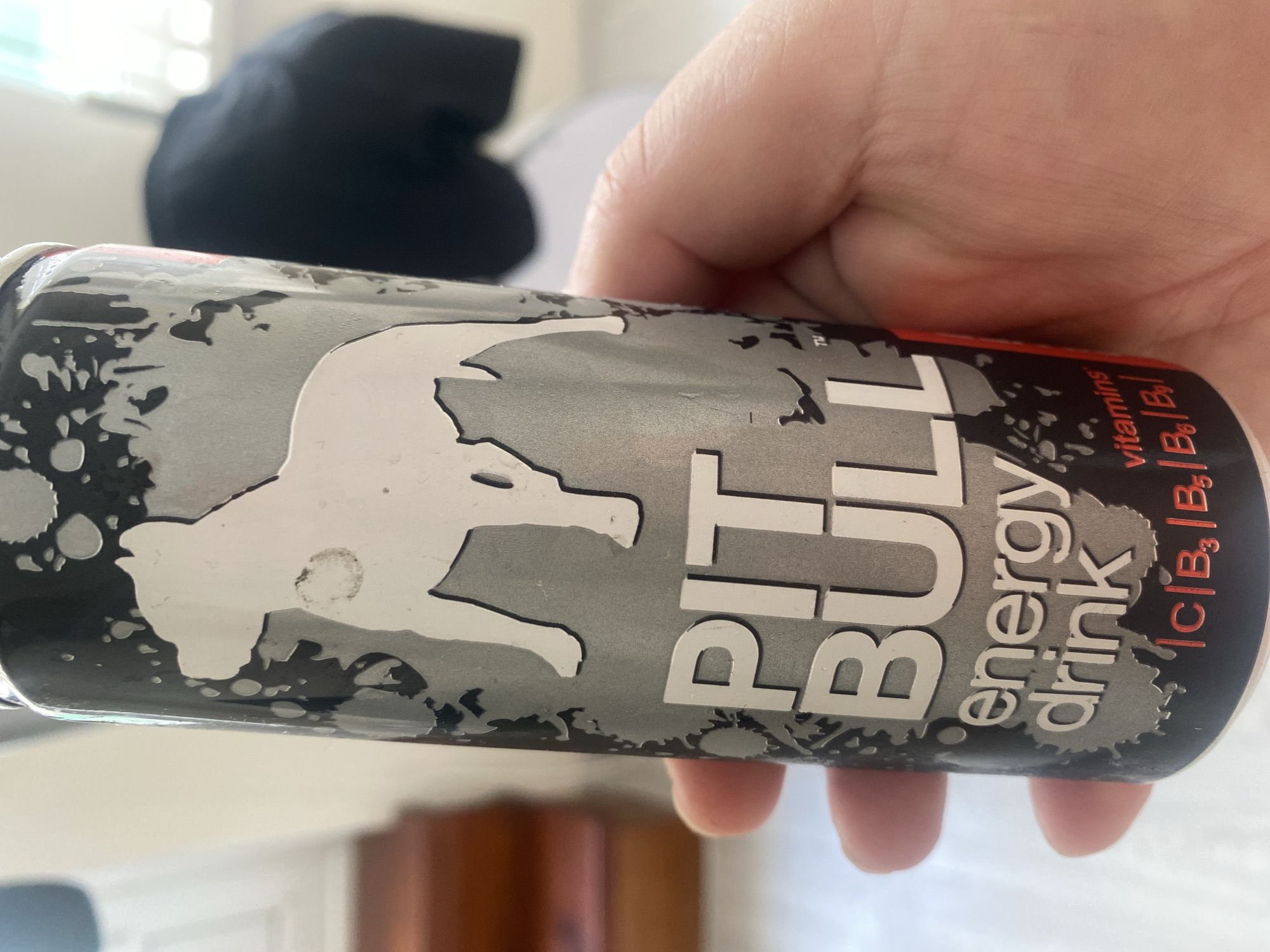 A person's hand holds a can of "PIT BULL energy drink." The can is primarily gray with a white silhouette of a dog. Below the dog, the text "PIT BULL" is prominently displayed in white. Further down, "energy drink" is written in a smaller font. At the bottom of the can, the word "vitamins" is visible, followed by a list of vitamin names: "C | B3 | B5 | B6 | B12". The background is out of focus but appears to be an indoor setting with natural light. There are no discernible landmarks or features to identify a specific location within Chișinău, Moldova.