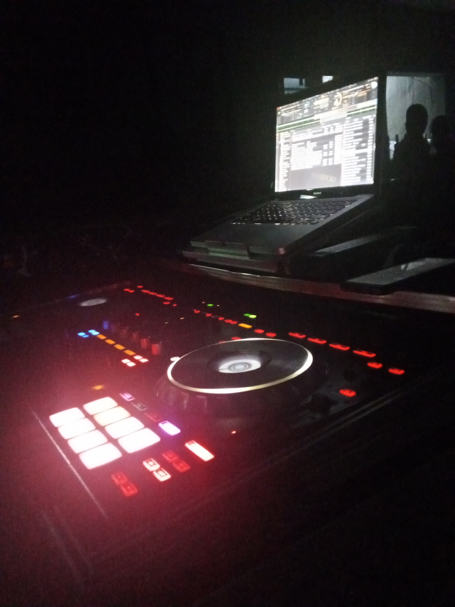 In a dark room, a DJ controller is illuminated by its own lights. A laptop sits on a stand behind the controller, its screen displaying audio software. Two silhouettes of people stand in the background to the right of the laptop. The location is Unknown, Unknown.