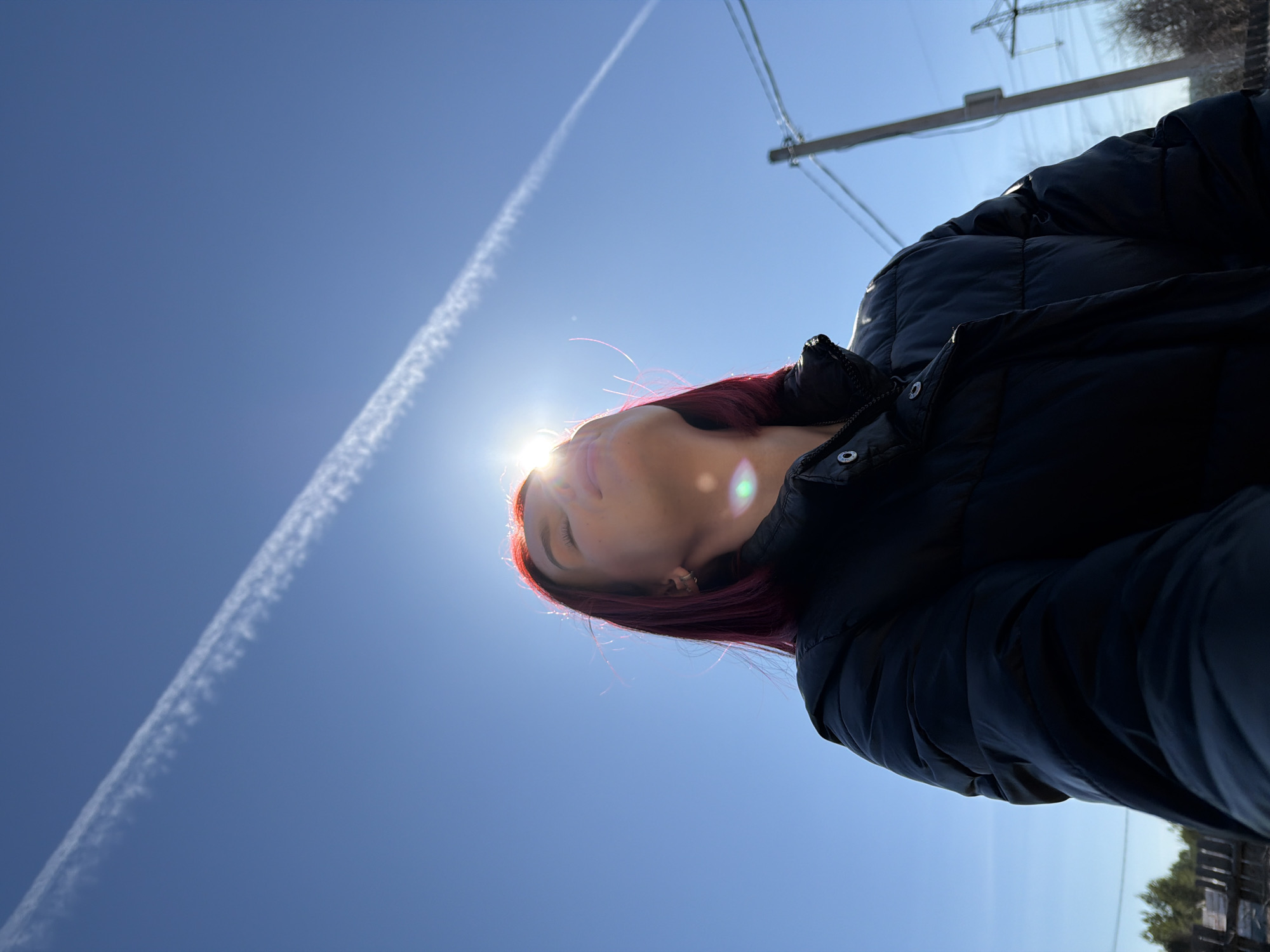 A woman with red hair is wearing a black puffer jacket. She is looking up towards the sun. A contrail is visible in the blue sky. A utility pole and wires are in the background. The location appears to be Chaikovsky, Russia.