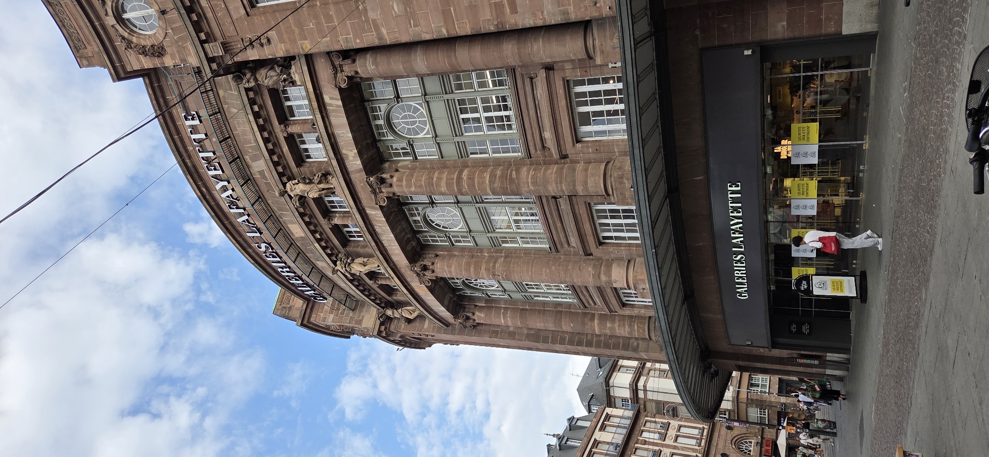 The image shows the exterior of the Galeries Lafayette department store in Strasbourg, France. The building is made of reddish-brown stone and features classical architectural elements, including columns and statues. The name "GALERIES LAFAYETTE" is prominently displayed in large, illuminated letters across the upper facade and above the entrance. The entrance area features a curved awning and large glass display windows. Several people are visible on the sidewalk in front of the store, some walking and others near display signs. The sky above is partly cloudy with a bright blue sky. The ground in front of the store is paved with cobblestones and there is a glimpse of a bicycle in the foreground.