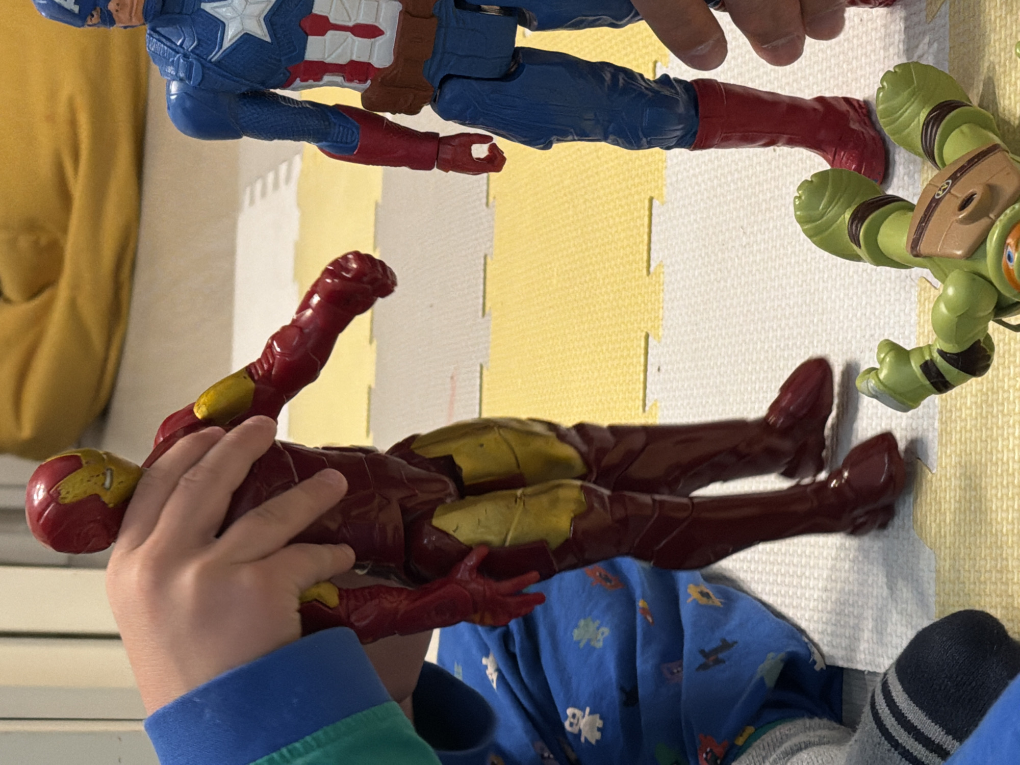 This image captures an indoor scene, likely a child's play area, with several action figures arranged on interlocking foam playmats. The playmat consists of alternating yellow and white square sections.
In the foreground, a child's hands are actively engaged in play, holding a red and gold Iron Man action figure. The child is wearing a blue and green long-sleeved shirt, and a blue and grey striped sock is visible towards the bottom right.
Three distinct action figures are present in the scene. A Captain America figure, clad in its iconic blue suit with red and white accents and a prominent star on the chest, is positioned towards the top. Its red boots are visible, pointing towards another figure. Lying on its back nearby is a light green Teenage Mutant Ninja Turtle figure, identifiable by its distinctive brown shell and green limbs. The Iron Man figure is held by the child, appearing ready for action.
The background includes a soft, yellow fabric, possibly part of a cushion or furniture. The setting suggests a casual, domestic environment, such as a living room or a child's bedroom, dedicated to playtime. No specific time of day, weather conditions, or emotional expressions are discernible. There is no visible text within the image. The provided location context for this activity is Sofiivska Borshchahivka, Ukraine.