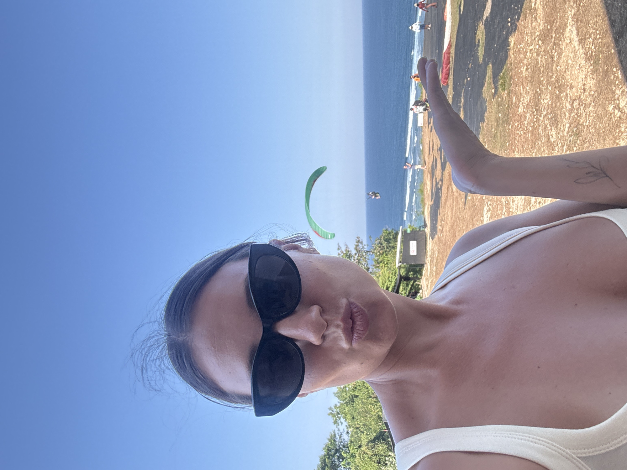 A woman with dark hair pulled back and wearing sunglasses and a white tank top is posing for a selfie with a paraglider in the background. She is puckering her lips and holding out her left hand. The paraglider is green and white, and a person is visible underneath it. The scene appears to be a cliff overlooking the ocean in Kuta Selatan, Indonesia, with several other people visible in the distance. The ground is covered in dirt and rocks. The sky is clear and blue.