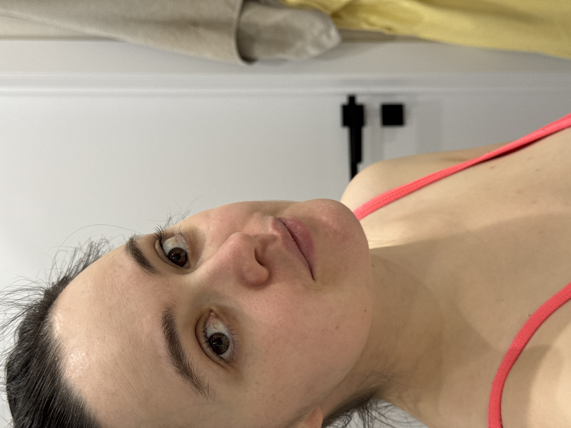 A close-up, angled shot of a woman with brown eyes and dark hair pulled back. She is wearing a pink athletic top. The background includes a white wall with a black object mounted on it, and above that, draped beige and yellow fabrics. There is no information available in the image to verify it was taken in Izhevsk, Russia.