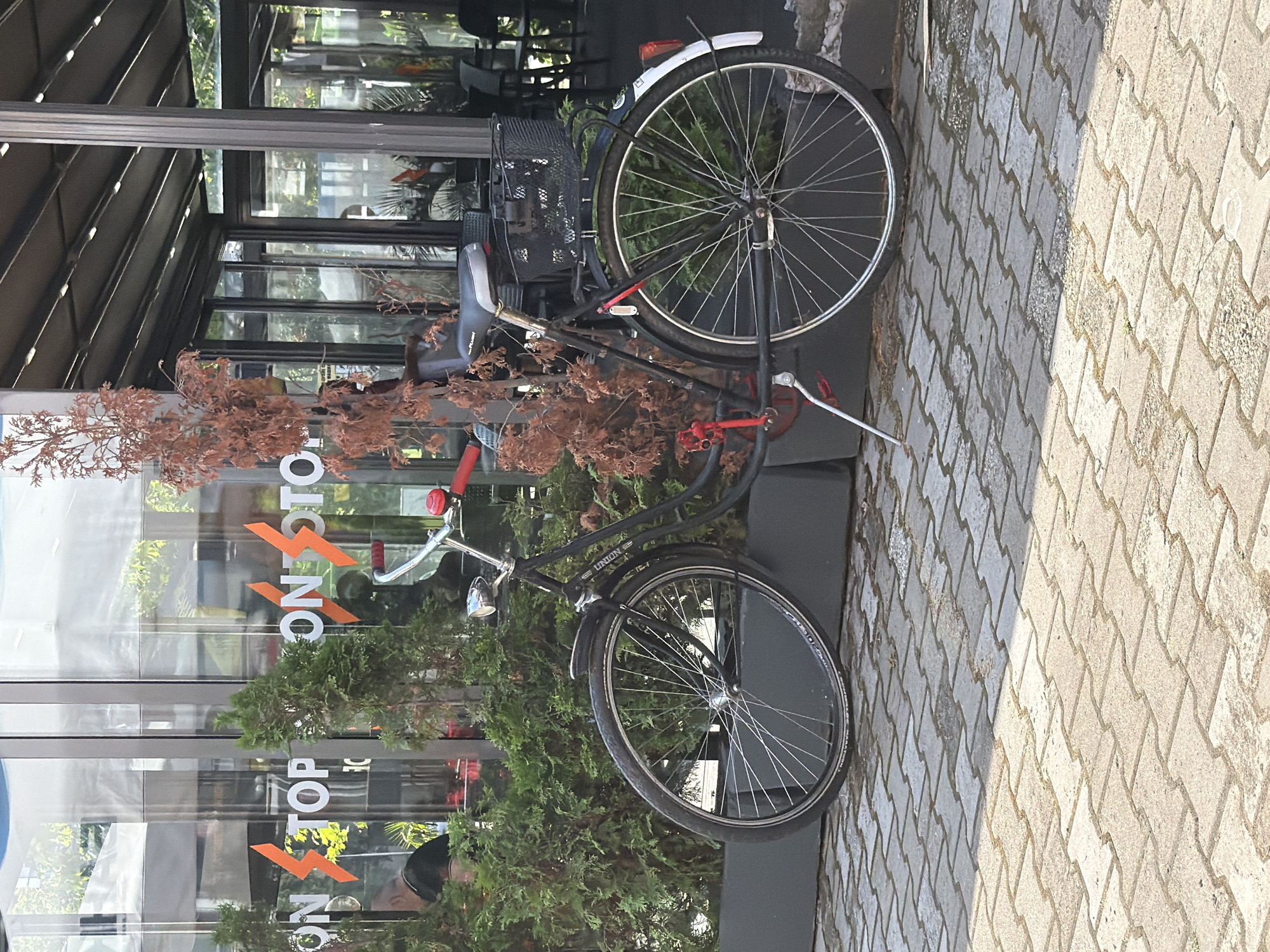 The image displays a black bicycle parked outdoors in Plovdiv, Bulgaria, beside a contemporary building with a glass facade. The bicycle, a "UNION" brand as indicated on its frame, features a front basket, white mudguards, a silver bell, and red accents on its handlebars and pedals. It is leaning against a dark planter or low wall, positioned amidst a mix of lush green bushes and dried, reddish-brown plant foliage.
The building's extensive glass windows reflect the surroundings, including trees and a faint, distorted reflection of a person's head wearing a cap. Visible on the glass are the white and orange letters spelling "ON STOP" or "NON STOP," accompanied by a stylized orange V-shaped graphic. Above the windows, a dark, slatted awning extends over the building's entrance.
The ground in the foreground is a paved walkway made of light-colored, interlocking concrete pavers. Bright sunlight illuminates the scene, creating noticeable shadows on the pavement, suggesting it is daytime with clear weather. No direct human activity is depicted; the scene is static, capturing a moment in an urban or commercial setting.