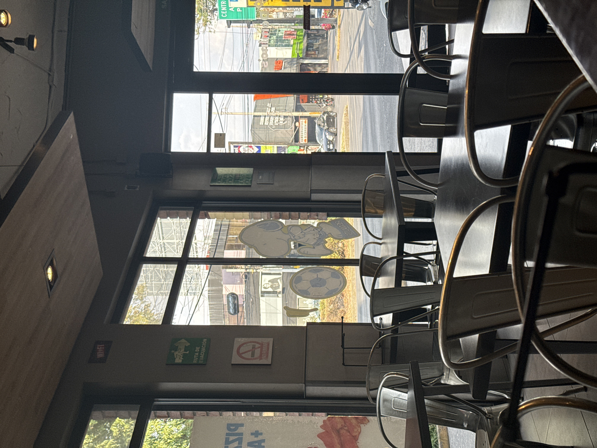 The interior of a restaurant in Puebla, Mexico is seen through a window. Metal chairs and tables are stacked and arranged. In the window facing the street there are decorations of Snoopy the cartoon character, a soccer ball, and other items. Signs for WIFI, a Route of Evacuation, and No Smoking are displayed. Outside is a sign that reads "Hamburguesas al carbon." A street sign for Central Avenida is visible.