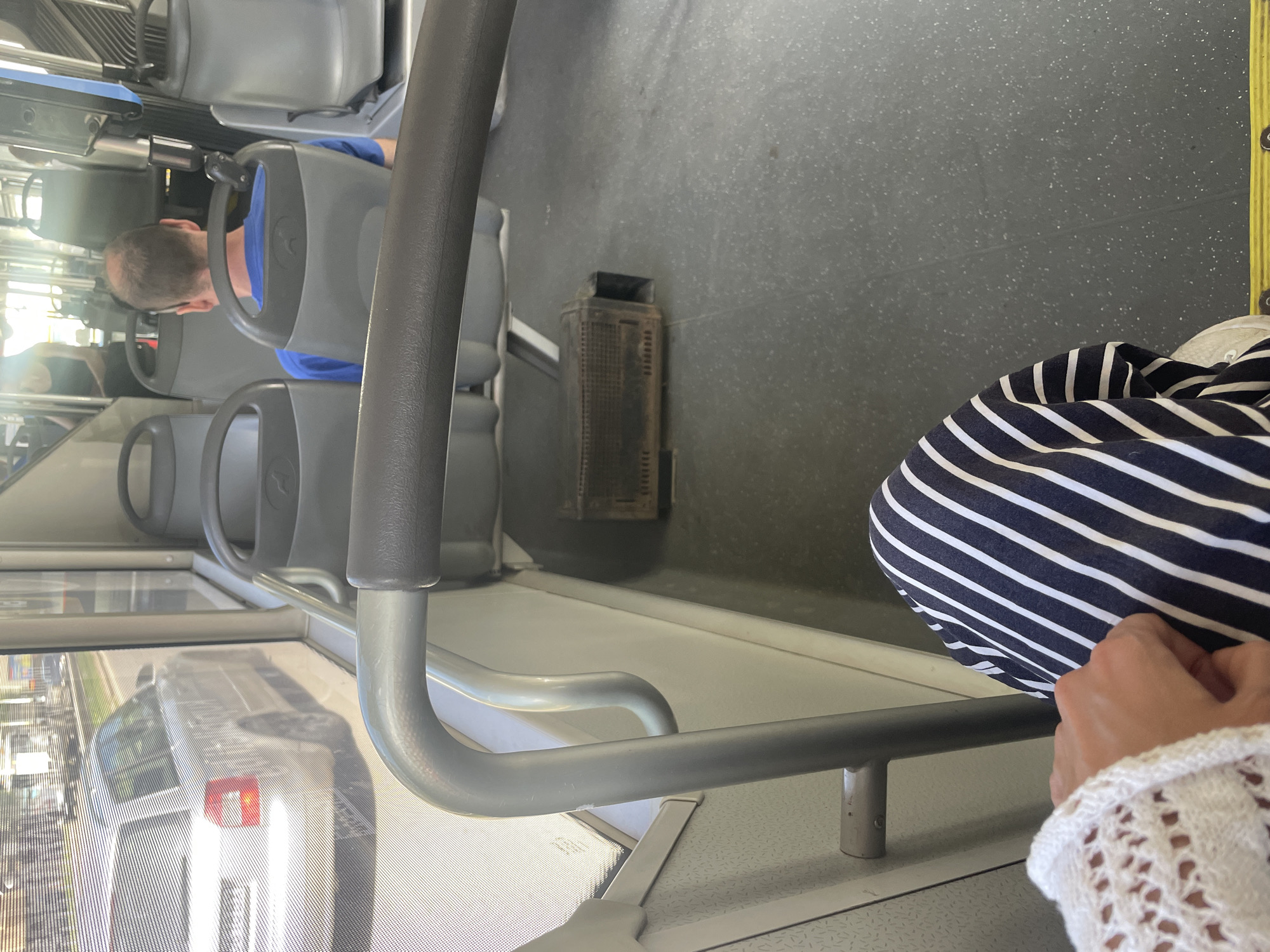 The image shows the interior of what appears to be a public transportation vehicle, likely a bus, with a view through the side window. The reflection visible in the window suggests a cityscape with cars and buildings, possibly taken in Sofia, Bulgaria. The vehicle's interior includes gray seats, a gray floor, and gray handrails. In the foreground, a person wearing a striped garment and a white lace sleeve is visible. Further back, another person with short, light-colored hair is seated. On the floor, there's a dark, rectangular object. The handrail is prominently in view, adding depth to the composition.