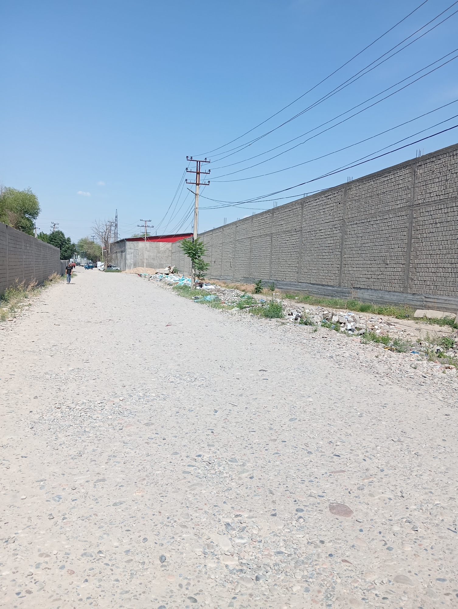 The image depicts a desolate dirt road in Eshanguzar, Uzbekistan. The road surface is composed of packed earth and gravel. On either side are high, unfinished gray brick walls. Litter and debris are scattered along the base of the walls. Power lines stretch across the sky above the road. In the distance, a person walks along the road. Several vehicles are also visible further down the road. The sky is clear and blue. A utility pole stands on the right side of the road.