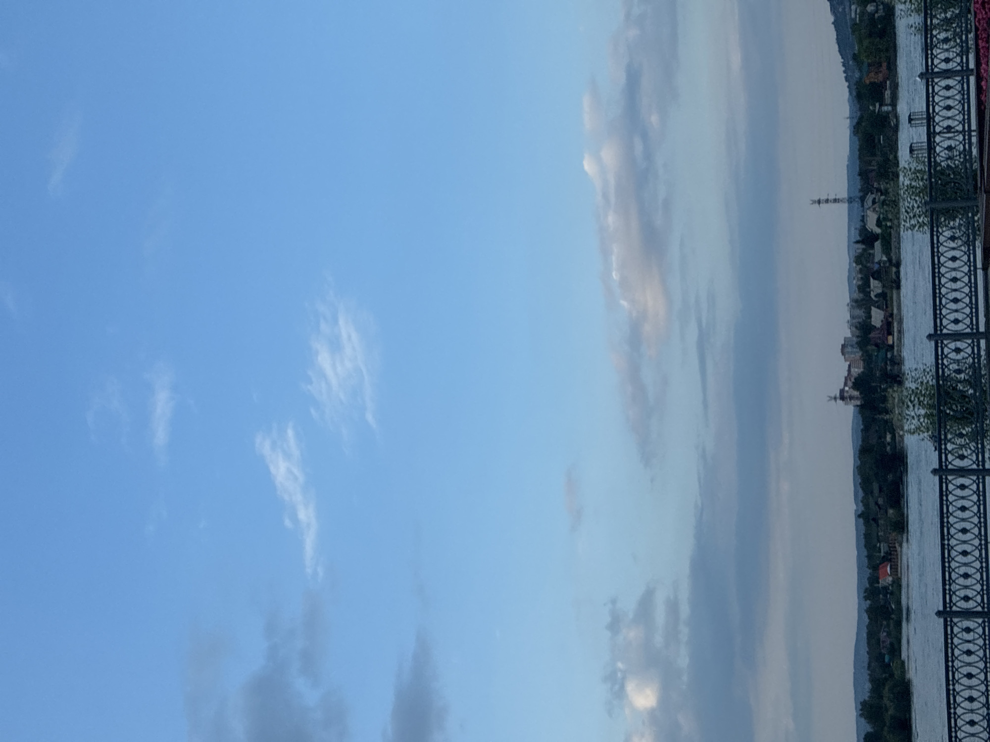The image shows a wide river with a city skyline in the background. The sky is a clear blue with some scattered white clouds. The city skyline consists of buildings, some of which appear to be residential, and a tall radio tower. There is a decorative metal fence in the foreground. The scene appears to be a waterfront view of Irkutsk, Russia.