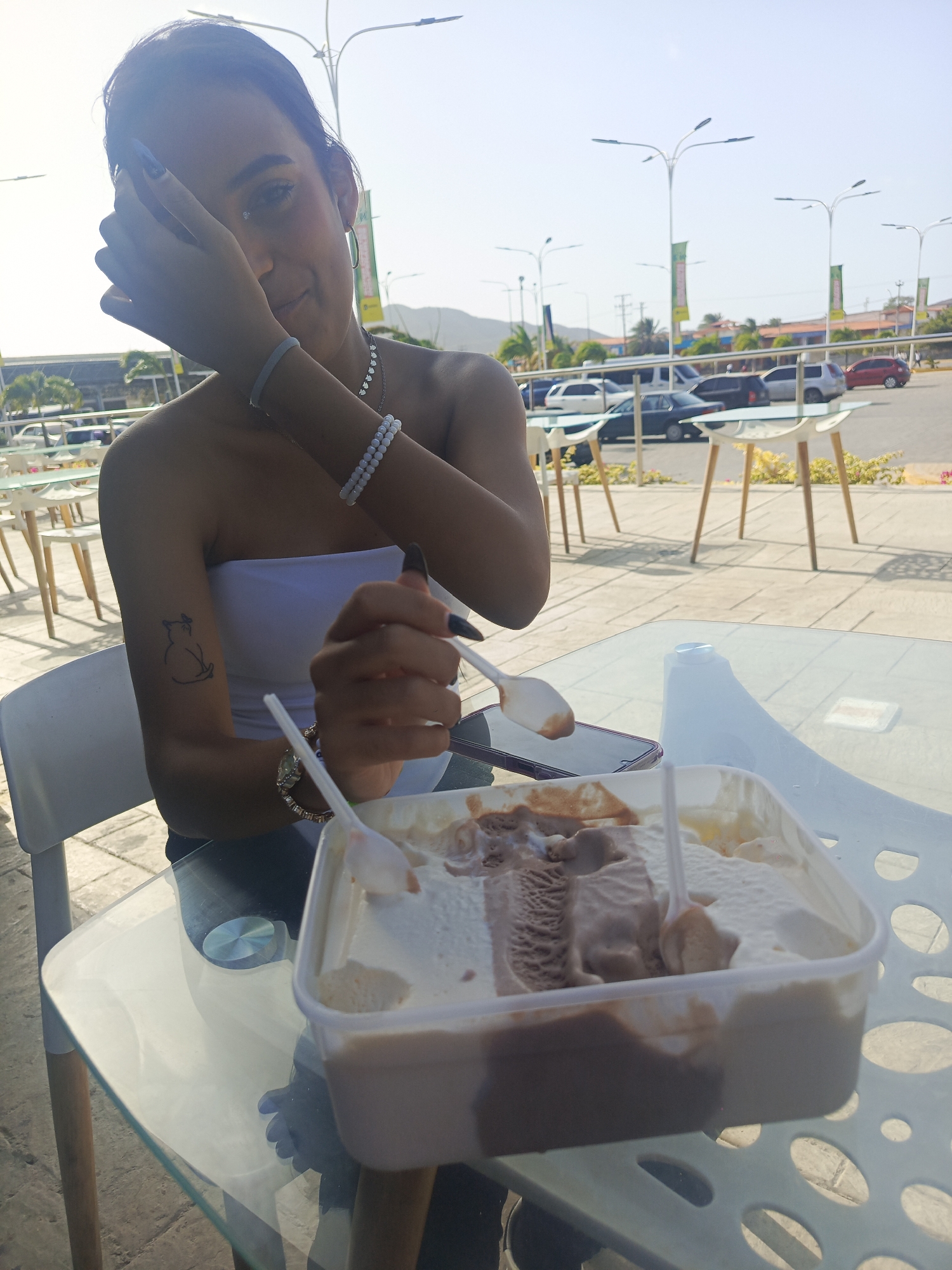 The image features a young woman enjoying a large container of ice cream outdoors on a bright, sunny day, likely in Juan Griego, Venezuela. She is seated at a glass-topped table with white chairs, holding a plastic spoon in her right hand and partially covering her face with her left hand, possibly shielding from the sun or expressing shyness, though a slight smile is visible around her left eye.
She wears a white strapless top, a silver necklace, a grey wristband, and two beaded white bracelets. A distinctive outline tattoo of a cat is visible on her right upper arm. Her fingernails are long and dark-colored. The ice cream, a mix of chocolate, vanilla, and possibly other flavors, is melting in a large rectangular plastic tub, with two plastic spoons in it. A smartphone is also on the table.
The setting is an outdoor seating area, likely part of a cafe or ice cream parlor. Beyond the dining area, there's a paved surface resembling a parking lot or street, with several cars parked or passing by. Modern, multi-armed street lamps with green banners line the street. In the background, under a clear, bright sky, are various buildings, palm trees, and distant hills or mountains. The strong sunlight and clear sky indicate it is daytime with warm weather. No legible text is visible in the image. The scene captures a relaxed, casual moment in a vibrant, sunny location.