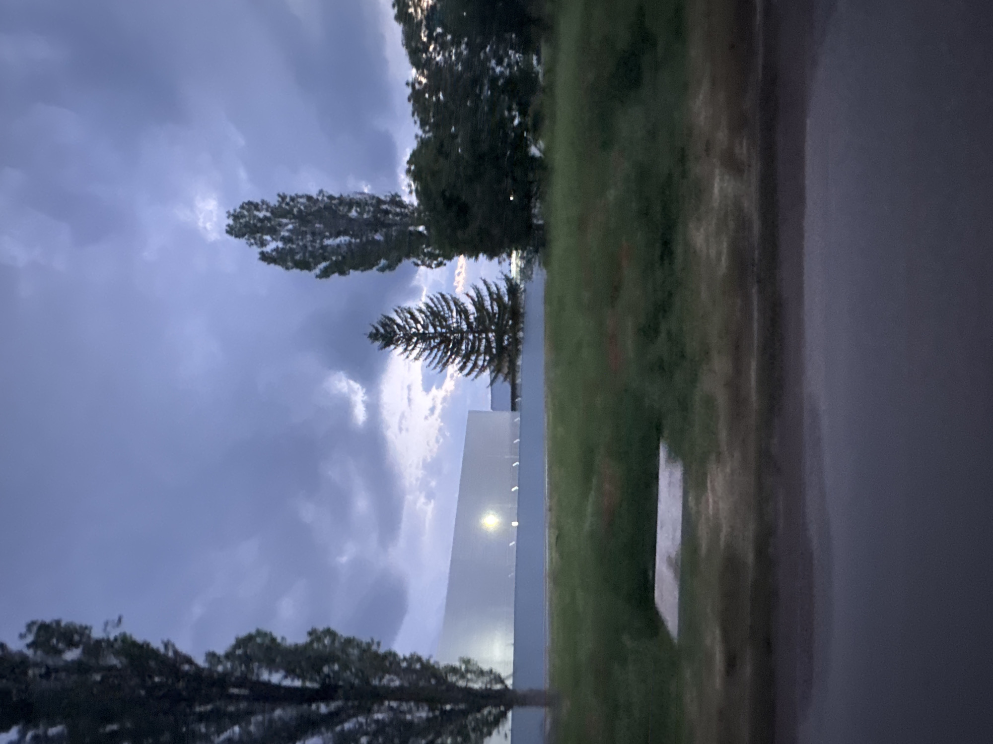 This image, taken in Yana, Bulgaria, depicts an outdoor scene during twilight or early evening. The sky, which occupies the upper half of the frame, is heavily overcast with dark grey and purplish clouds, though hints of brighter clouds suggest a fading light source.
In the midground, a prominent, light-colored, angular building or wall stands, featuring several artificial lights along its side. One of these lights is particularly bright, casting a strong, almost sun-like glare. Directly in front of and below this structure is a body of calm water, reflecting the building's lights and the surrounding trees, though the reflection is somewhat indistinct.
Various trees are silhouetted against the dim sky and reflected in the water. A tall, slender conifer stands out to the left of the building, alongside other broader, leafy trees scattered across the scene. A strip of green grass runs vertically through the right side of the image, bordering a dark, paved surface that appears to be a road or pathway. The overall atmosphere is quiet and subdued, characteristic of dusk with artificial illumination beginning to dominate. There are no visible people, specific activities, or text within the image.