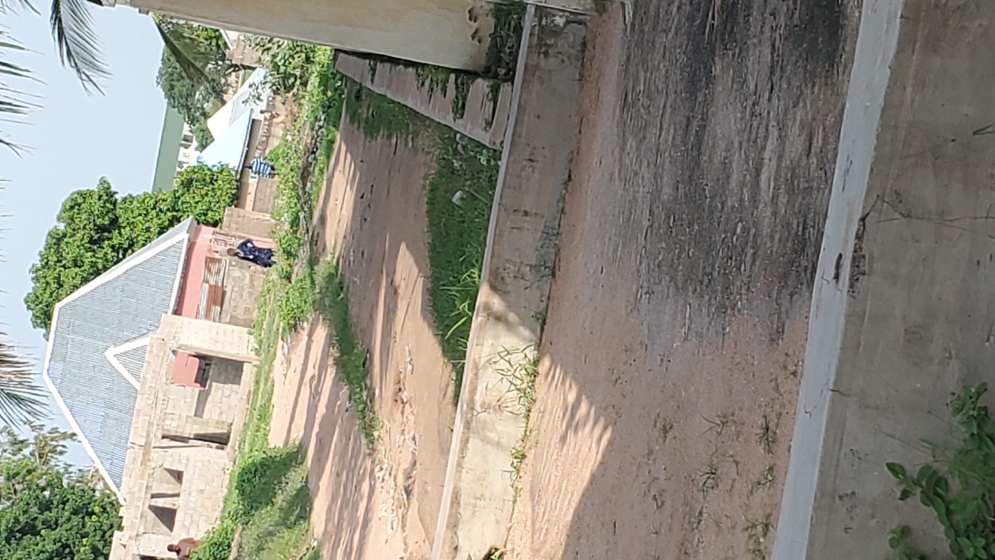 The image shows an outdoor scene in Jalingo, Nigeria, likely a residential area under construction or development. In the foreground, there is a dirt path leading towards several buildings. To the left, a partially constructed building made of concrete blocks is visible, with a person standing near it. Further back, a finished building with a corrugated metal roof is prominent, painted in pink and white. To the right of this, another building with a light blue roof is visible. In the middle ground, a person in a dark blue garment is standing near a wall. Another person in a striped shirt is walking further into the scene. The environment is characterized by dry, sandy ground, patches of green grass, and scattered debris. Trees are visible in the background, suggesting a warm climate. The scene appears to be in daylight.