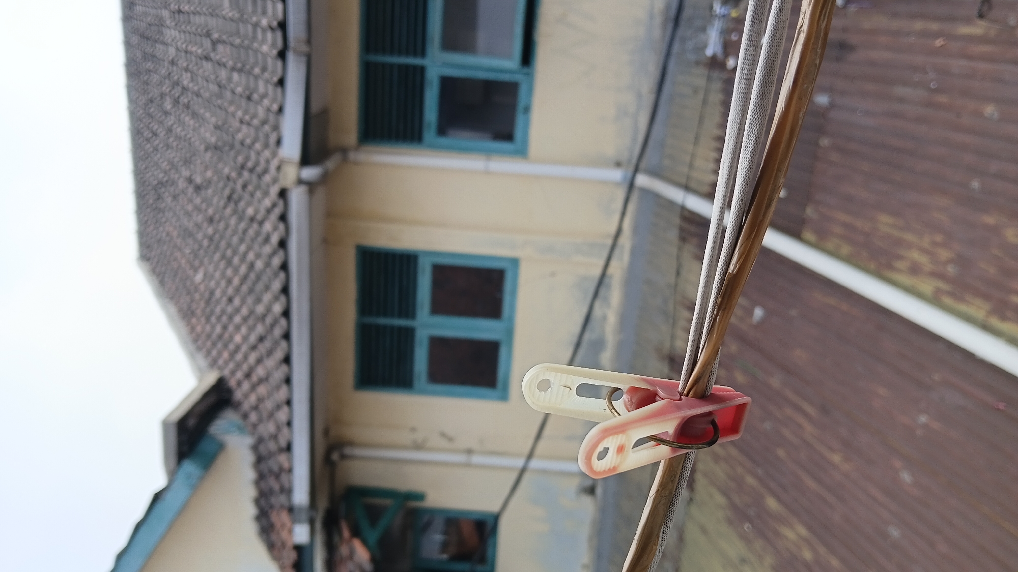 The image shows a clothesline in front of a yellow building with a tiled roof. There are two windows with turquoise frames and louvred shutters. A red and white clothespin is holding some strings. The background is slightly blurred, showing a tin roof. The overall impression is of a residential building in an urban setting. This scene could be in Bandung City, Indonesia, as the architectural style and materials are common in the region.