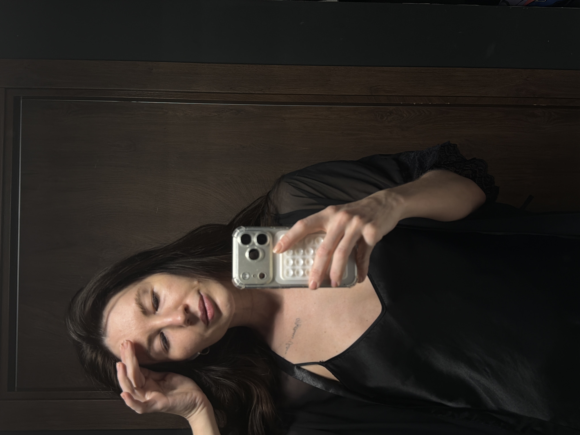 A woman is taking a selfie indoors. She is wearing a black robe with lace detailing. She is holding a cell phone in her right hand. She has dark hair. Her left hand is touching her forehead. A tattoo is visible on her chest. A brown door is visible in the background. The city of Perm, Russia, is not discernible from the visual information provided.