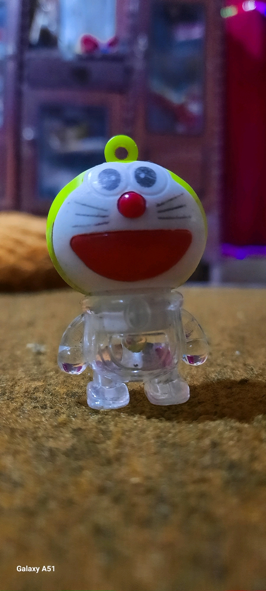 A close-up, low-angle, macro shot shows a transparent plastic toy figure, resembling the cartoon character Doraemon, placed on a textured brown surface. The toy has a prominent round head with large, black eyes, a red nose, and a wide red mouth. A small green loop extends from the top of its head. The body of the toy is clear plastic, allowing a view of internal colorful elements. The background is blurred, featuring hints of furniture and colorful lights. The bottom left corner of the image displays the text "Galaxy A51". There is no visible indication of the city of Jatitengah, Indonesia, in the image.