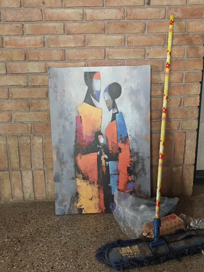 A vertical photograph captures an indoor or covered outdoor scene in Kalyan-Dombivli, India. A large, rectangular abstract canvas painting, measuring approximately 1.2 meters in height, leans against a light brown brick wall. The painting depicts three stylized, faceless figures: a taller figure on the left in yellow and orange hues, a slightly shorter figure on the right in orange and blue, and a smaller figure centrally positioned between them. The painting's background is textured grey. To the right of the painting, a blue flat-head mop, approximately 1.5 meters tall, leans against the same brick wall. Its handle is yellow with a pattern of small red and white flowers, and its dark blue mop head rests on the floor. The floor consists of dark grey speckled terrazzo or polished concrete. Beneath the painting and mop, on the floor, are a crumpled transparent plastic bag and a partially visible brown paper bag containing unknown items, accompanied by other indistinct debris. No individuals are present in the scene.