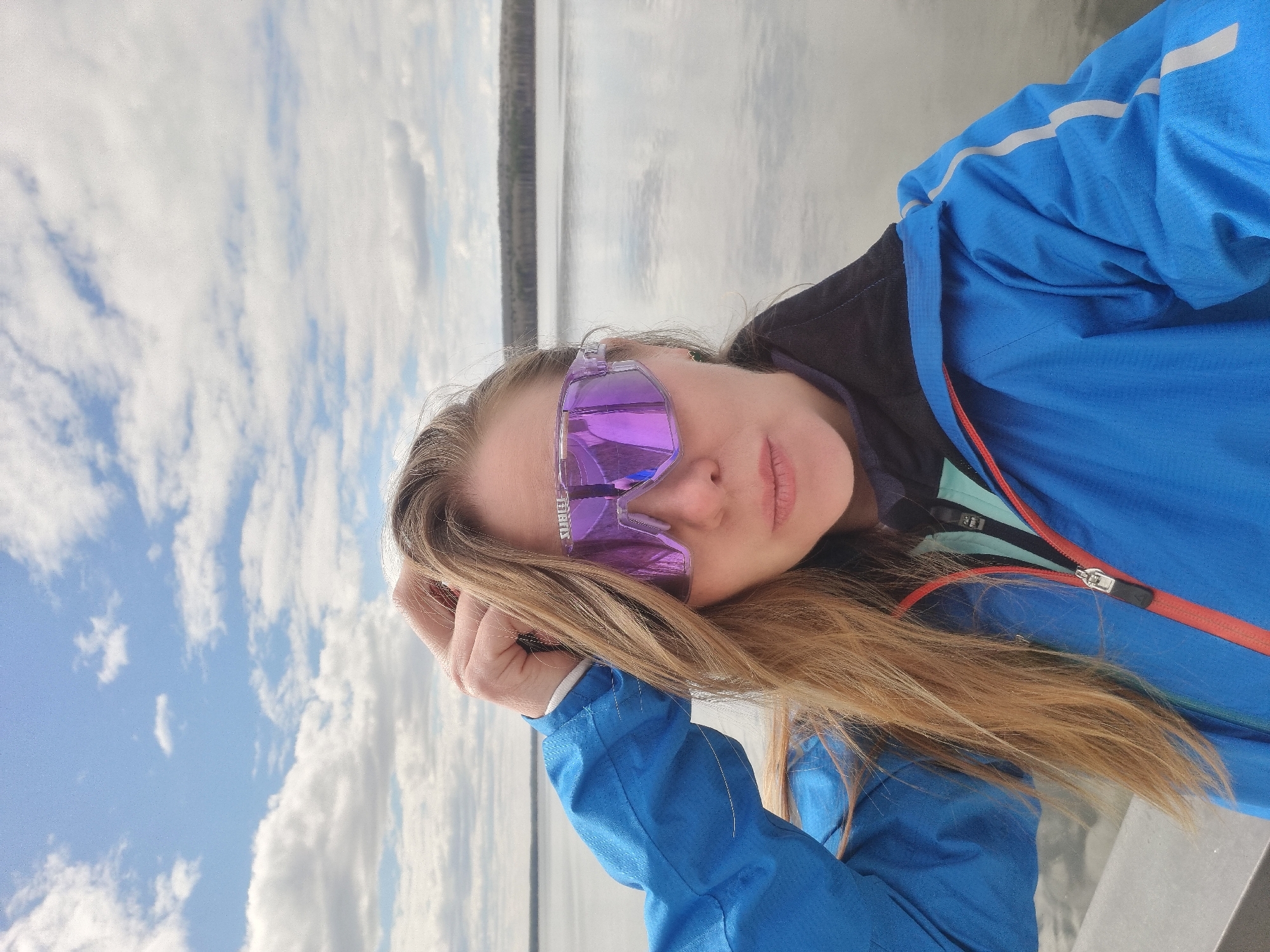 A person wearing purple sunglasses and a blue jacket is posing for a selfie outdoors. The background features a body of water and a sky with clouds. The location is Chaikovsky, Russia.