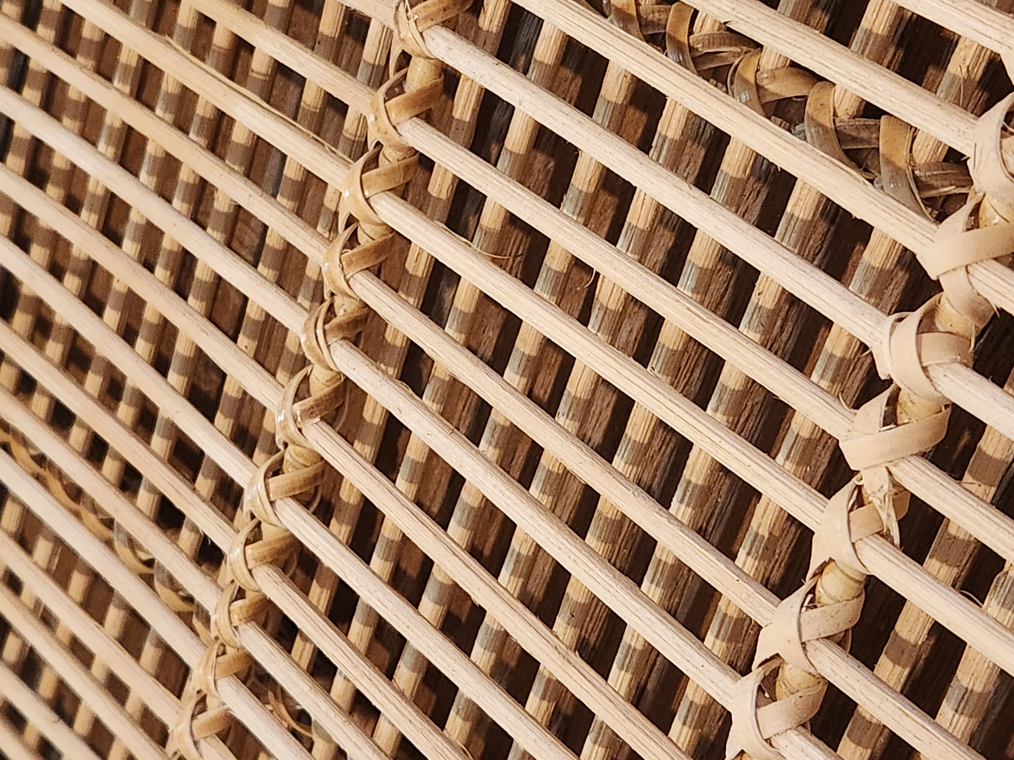 This media file shows a close-up, diagonal view of a woven bamboo or rattan surface. The material is light brown and appears to be a natural reed or grass. The weaving creates a grid pattern, with thin, rounded strands running parallel and diagonally across each other, forming small square openings. In some areas, thicker, lighter-colored strips of woven material are used to secure the grid structure, creating a decorative knot-like effect. The background visible through the openings is a darker, textured brown, suggesting an underlying surface or object. The lighting appears to be natural, casting shadows that highlight the texture and depth of the weave. The overall impression is one of rustic craftsmanship and natural materials. There is no visible text, people, or indication of a specific time of day or weather. The location context provided is Maitland, United States of America, which suggests this could be part of furniture, a decorative screen, or architectural element.