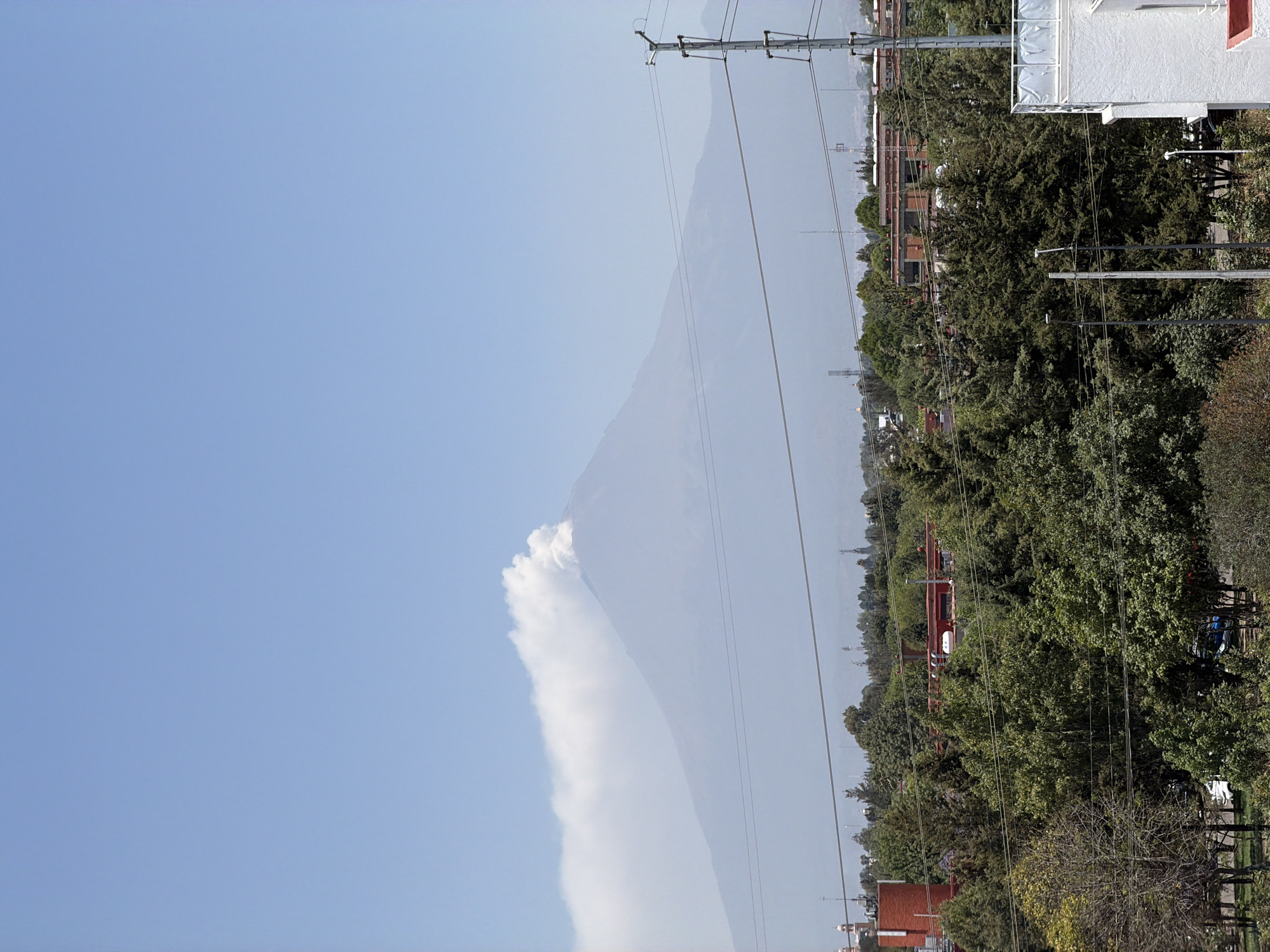 The image displays a panoramic view of an urban landscape under a clear, bright blue sky, dominated by the distant Popocatépetl volcano, identified in the context of San Andrés Cholula, Mexico. A prominent white plume of smoke and ash billows from the volcano's peak, extending horizontally across the midground to the left. The volcano itself is a large, hazy gray mass, its conical shape subtly defined against the sky.
In the midground and foreground, a dense canopy of lush green trees fills the lower half of the scene, indicating a vibrant, possibly residential or park-like area. Scattered among the trees are various buildings, some with red-tiled roofs, suggesting a blend of traditional and modern architecture. On the far right, a large white building features a prominent, blank billboard-like structure on its roof.
Multiple tall utility poles with numerous power lines crisscross the upper-mid section of the image, creating a network of wires against the sky and above the treeline. The scene is illuminated by bright daylight, suggesting a clear morning or afternoon. No specific people or discernible text are visible. The overall impression is one of daily life unfolding amidst the powerful and active presence of a natural wonder.