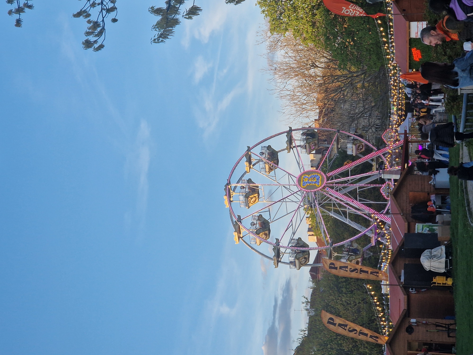 This media file captures an outdoor fair, market, or festival scene during the late afternoon or early evening. The image is dominated by a brightly lit Ferris wheel positioned in the mid-ground, featuring multiple gondolas and illuminated with pink and white lights along its frame. The center hub of the Ferris wheel clearly displays the word "FUN" in a stylized font.
The sky above is a clear, vivid blue with faint wispy clouds, suggesting pleasant weather. Trees with both verdant green leaves and bare branches frame the upper and right sides of the scene, indicating either a transitional season or mixed flora.
The foreground and right side of the image reveal a lively gathering. Numerous people are present, strolling, conversing, and possibly dining at partially visible tables. Several wooden stalls or booths, adorned with warm, glowing string lights, line what appears to be a pathway. These lights are illuminated, suggesting the time is approaching dusk or has just entered the early evening. A man with a beard and an orange jacket is visible among the crowd on the right side.
Prominent text on various banners identifies some of the offerings: a large orange banner reads "APEROL SPRITZ," while two smaller, beige banners proudly display "PASTA." The overall ambiance is festive and social, with people enjoying the outdoor event.