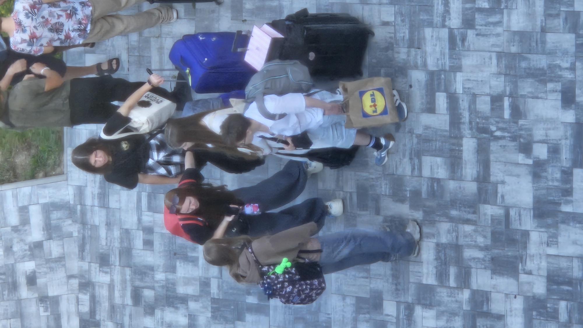 The image, taken from a high angle, shows a group of young people gathered on a paved outdoor area. The paving stones are a mix of grey tones with a textured, wood-like pattern. To the right of the frame, a patch of green grass is visible. The scene appears to be a public space, possibly a pedestrian zone or a plaza.
There are at least six individuals visible. One person, wearing a black t-shirt and a plaid shirt tied around their waist, is looking down and holding a light-colored tote bag. Another individual in the foreground is wearing a brown hoodie and jeans, with a backpack adorned with a colorful pattern and a green charm. A person in a white shirt and light-colored shorts is holding a brown paper bag with a "Lidl" logo and carrying a backpack. Another person, wearing dark pants and a red backpack, is bent over. Two other individuals are also present, one in a white top and the other in dark clothing.
Various pieces of luggage are scattered around the group. These include a bright blue suitcase, a black rolling suitcase, and a pink and white suitcase. One person is holding onto a metal pole that appears to be attached to a wheeled object, possibly luggage or a stroller.
The individuals' clothing suggests a casual, possibly warm-weather setting. Their postures and the presence of luggage indicate they might be preparing for travel, waiting for transportation, or have just arrived. The overall impression is a snapshot of a group of young people with their belongings in an urban environment. The specific location in Prešov, Slovakia, cannot be determined from the visual cues in the image alone.