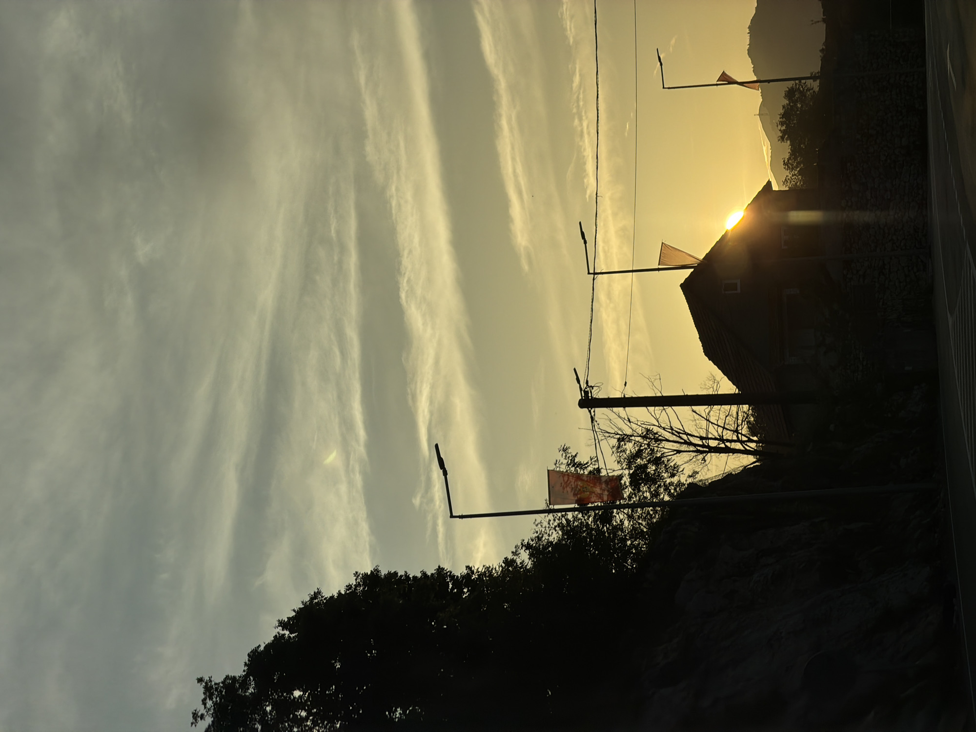 The image depicts a twilight scene in Crna Greda, Montenegro. The sky is filled with long, striated clouds. The setting sun is partially obscured by a dark, silhouetted building with a gabled roof, creating a sunburst effect. The silhouette of a mountainous landscape is visible in the background. In the foreground, there are trees and rock formations. Power lines and streetlights are present. A flag with a design is attached to one of the streetlights.
