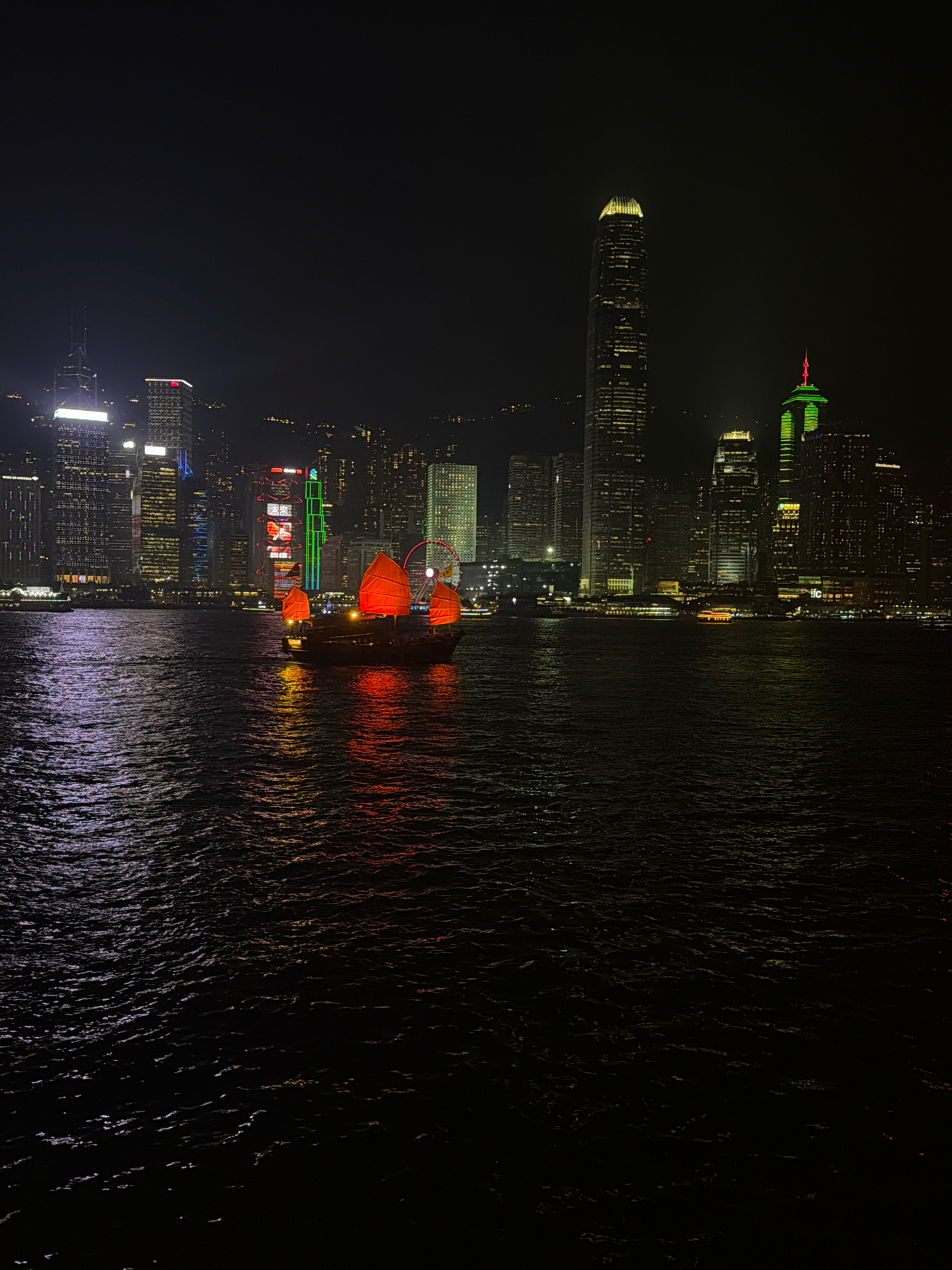 An eye-level, full shot captures the Hong Kong skyline at night, viewed from the water. In the foreground, a traditional Chinese junk boat with bright red sails glides across dark, rippling water that reflects the city's lights. Behind the boat, the Hong Kong skyline is illuminated by a myriad of building lights. Prominent skyscrapers, including the distinctive IFC Two building with its green top, and the Bank of China Tower with its pointed spire, are visible. The overall scene is a vibrant display of urban illumination against the darkness of the night sky and sea.