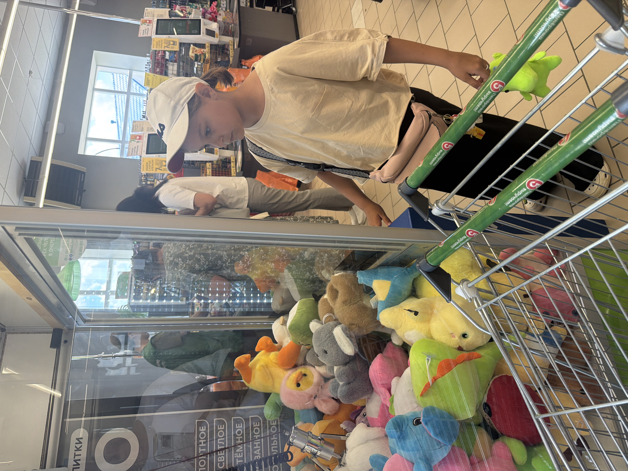 In Leningradskaya, Russia, a young girl wearing a white baseball cap and a beige t-shirt stands in front of a claw machine filled with plush toys. She is looking intently at the toys inside the machine. The claw machine is situated within a brightly lit store, likely a supermarket, as evidenced by the shopping carts and aisles visible in the background. The girl has a pink fanny pack slung across her body. Other shoppers and store displays are present in the background, indicating a public space. The floor is tiled, and overhead lighting illuminates the scene.