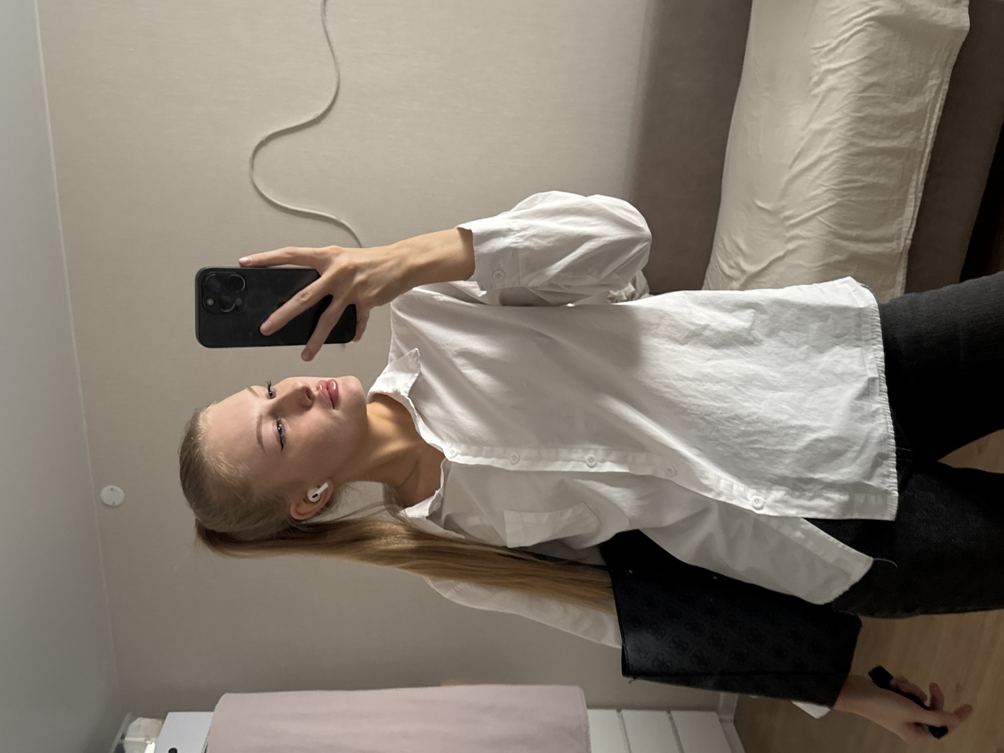 A young Caucasian female with blonde hair in a high ponytail is taking a selfie in a room. She is wearing a white collared button-up shirt and dark pants. She has white earbuds in her ears and is holding a black smartphone in her right hand, which she is using to take the photo. A black tote bag is slung over her left arm. The background shows a light-colored wall with a cord running along it, a glimpse of furniture, and a white cabinet. The floor is wooden. The image was captured indoors in Perm, Russia.