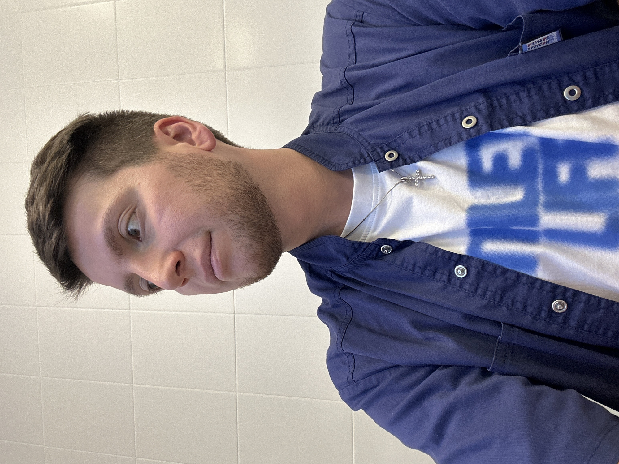 A young man with short brown hair and stubble is captured in a close-up, slightly angled shot. He is wearing a white t-shirt with a bold blue graphic design and a dark blue collared shirt over it. A silver cross necklace is visible around his neck. The man is looking off to the side with a slight smile and an expression that could be interpreted as amused or intrigued.
The background consists of a tiled wall, suggesting an indoor setting, possibly a bathroom or a changing room due to the lighting and tile pattern. The lighting is bright and even, indicating it's likely daytime or under artificial lighting. No specific time of day or weather can be inferred.
A small label on the inside of the blue shirt is visible, with the text "MEDICAL SERVICE" printed on it. The overall impression is a casual and candid shot, focusing on the subject's facial expression and attire. The location context of Moscow, Russia, does not provide any direct visual cues in the image itself.