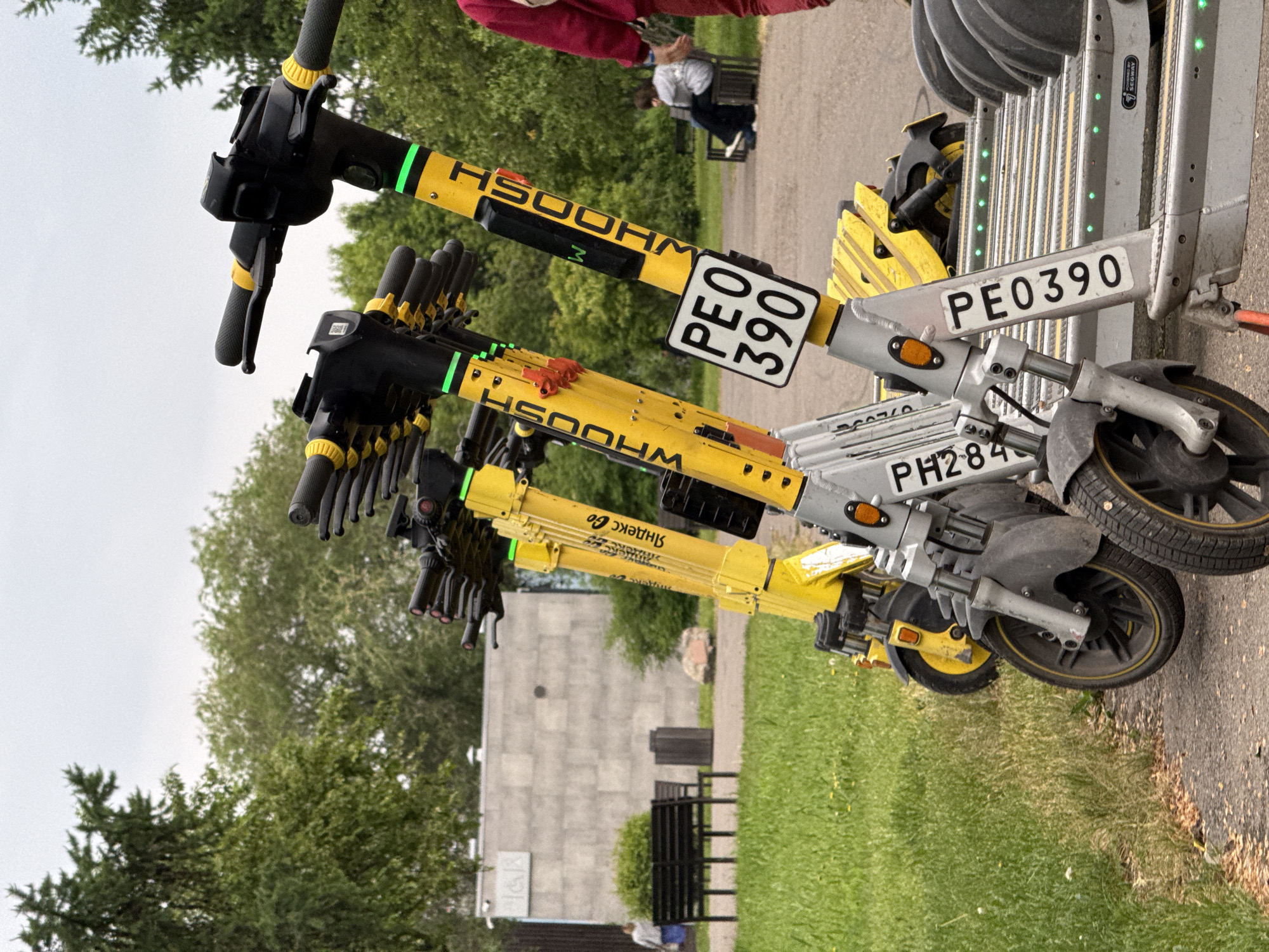In Krasnoyarsk, Russia, a row of yellow and silver electric scooters is parked on a paved path next to a grassy area. The scooters have "WHOOSH" branding and license plates. In the background, a building with a gray, textured facade and a bench are visible. Two people are seated on the bench, and another person is walking away from the camera on the left. The sky is overcast.