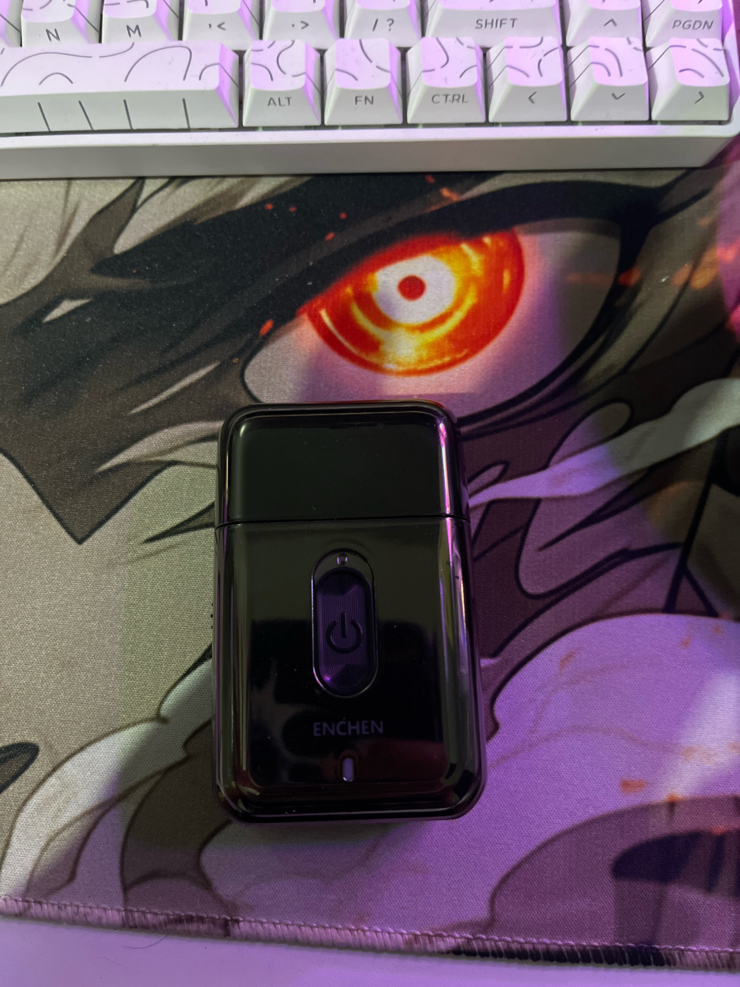 A black electric shaver with a purple power button is positioned on a mousepad with an anime-style eye design. The mousepad is placed on a surface below a white keyboard. The lighting in the image is purplish. The location is not specified in Cimahi, Indonesia.