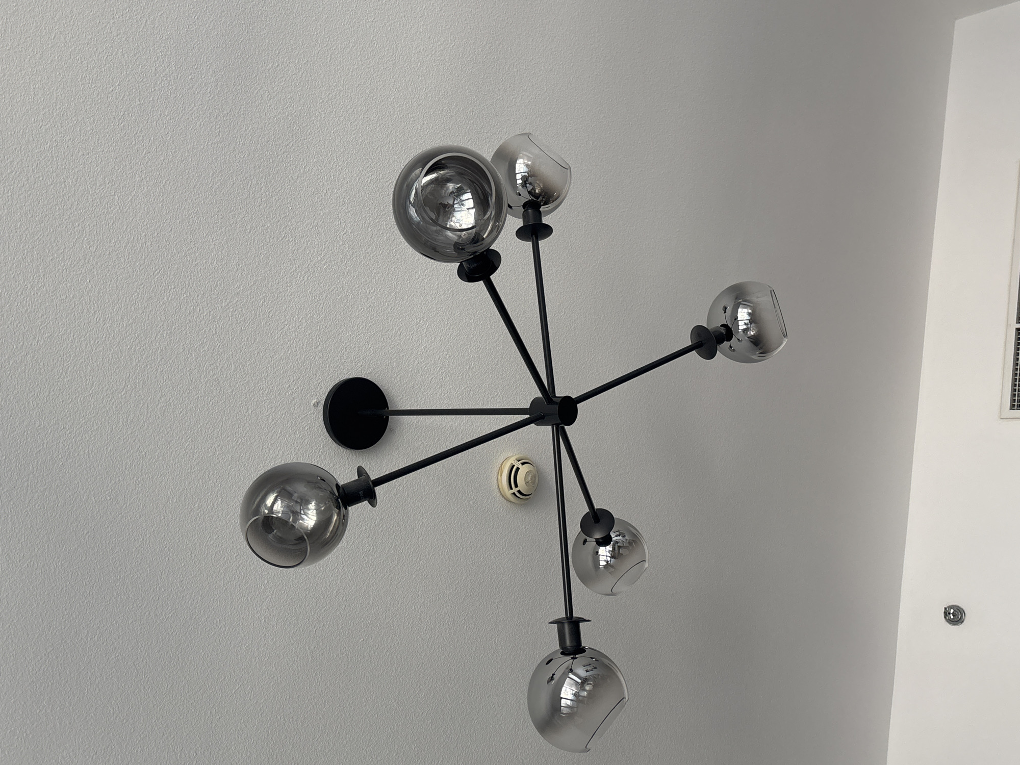 The image depicts a modern, black Sputnik-style chandelier with six smoked glass globes. The chandelier is mounted on a textured, white ceiling. A small, circular, beige smoke detector is visible to the right of the chandelier's central mounting point. The lighting in the image is diffuse, suggesting an indoor environment with natural or ambient light. The overall scene is clean and uncluttered, indicative of a contemporary interior space. The image does not provide any specific geographical context to determine if it is located in Dubai, United Arab Emirates.