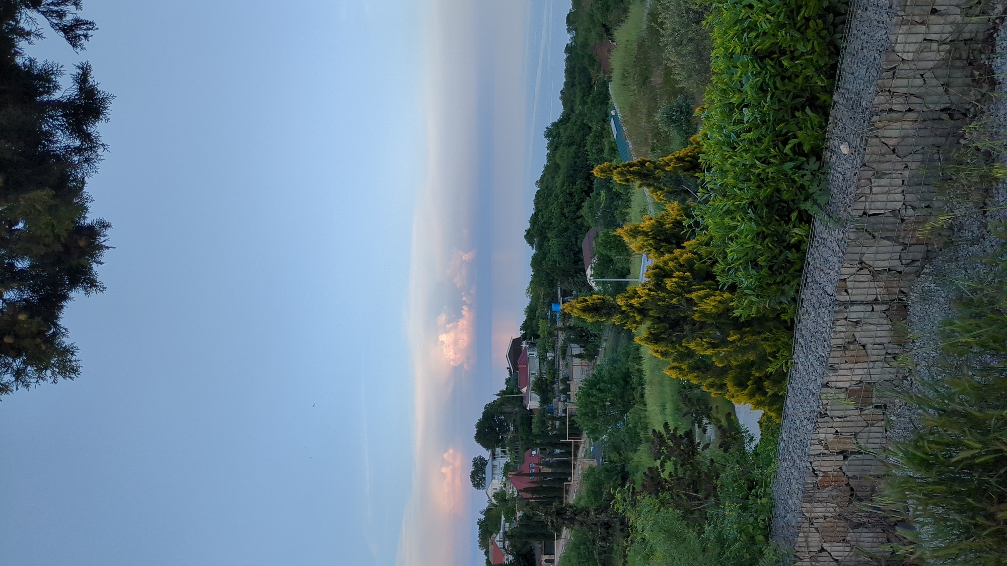 The image depicts a view of Kiparisnoe, Ukraine, possibly taken from an elevated position. The foreground features greenery, including trees and shrubs, with a stone wall retaining the landscape on the right side. Beyond the vegetation, there's a view of residential buildings with visible rooftops and surrounding trees. In the distance, the sky is partially cloudy with varying shades of blue and hints of pink, suggesting either sunrise or sunset. The composition gives a sense of looking out over the town towards the horizon.