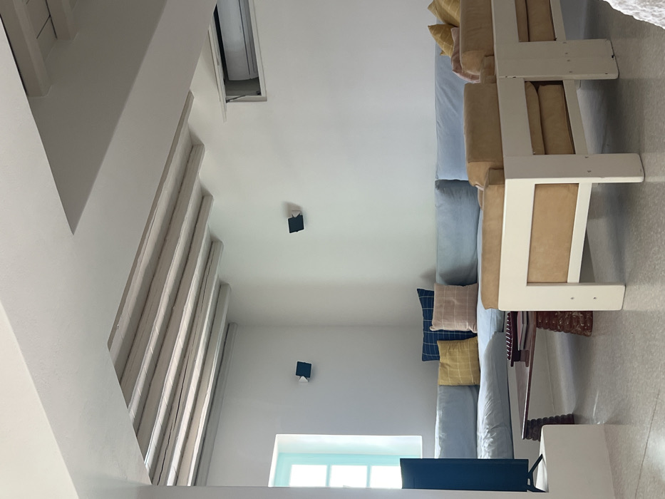 The image displays an unoccupied living room located in Tourlos, Greece. The room features white walls and a light-colored, flecked floor. The ceiling is white, with a section revealing exposed, horizontally arranged wooden beams painted a light color. A light blue-green framed window is positioned on the left wall, admitting natural light. Two dark, geometric wall-mounted light fixtures are visible, one on the left wall and another on the far wall. A white air conditioning unit is recessed into the upper right portion of the far wall.
An L-shaped sectional sofa with light blue cushions occupies the corner of the room, extending along the left and back walls. Several patterned throw pillows in yellow, dark blue, and light pink/peach colors are arranged on the sofa. In front of the sectional, a low, dark wooden coffee table with carved legs is present, bearing a dark, rectangular decorative object. To the right, two individual armchairs with white frames and light brown cushions are partially visible in the foreground. A dark flat-screen television is situated on a low, white built-in stand or shelf near the window on the far left. No individuals or active interactions are depicted.