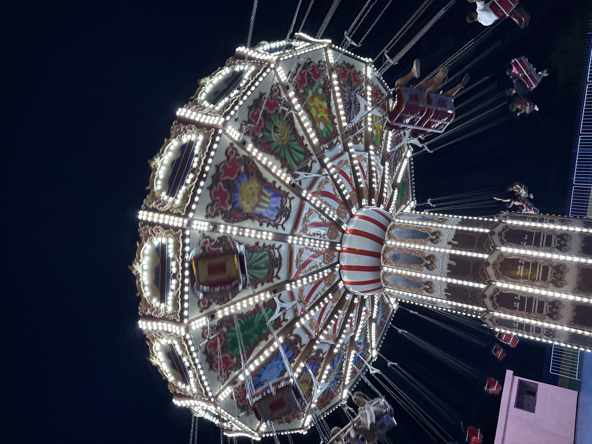This image captures a brightly lit carousel ride at night, likely in Bodrum, Turkey, given the context provided. The carousel is adorned with numerous small lights and decorative panels featuring colorful illustrations, including a cupcake and various abstract designs. People are seen seated in the swinging chairs of the ride, some appearing blurred due to motion. The dark night sky serves as the backdrop, contrasting with the illuminated ride. In the lower left corner, a small pink structure with a window is visible, and a white fence runs along the bottom of the frame. The scene conveys a festive atmosphere typical of an amusement park or fairground.