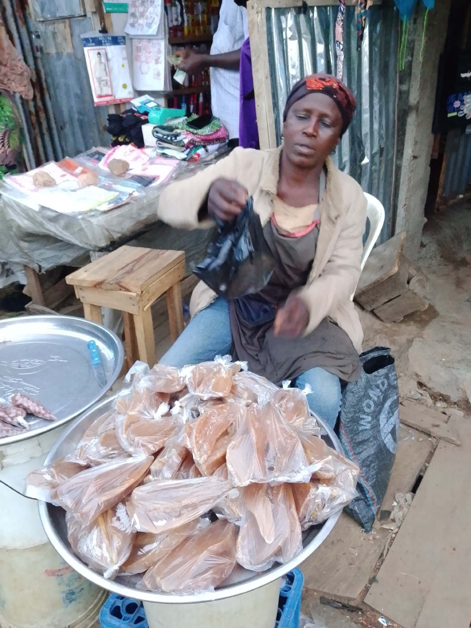 A woman, wearing a headscarf and a light-colored jacket over an apron, sits in a chair at an outdoor market stall. She is holding a black plastic bag in her right hand, appearing to be in the process of handing it over or preparing it. In front of her is a large metal bowl filled with numerous items individually wrapped in clear plastic, which appear to be food products, possibly candy or sweets, given their translucent, amber color. To the left of this bowl, another metal tray contains what look like sausages or some kind of cured meat. Behind the woman, the stall is set up with shelves displaying various goods, including folded clothing items like underwear and socks, and some packaged products. A small wooden stool is visible to her left. The background shows a corrugated metal wall, suggesting a rustic or informal market setting. The lighting indicates it could be daytime, but the specific time or weather conditions are not discernible. The woman's facial expression is neutral, and her posture suggests she is engaged in a transaction or preparing for one. There is no visible text in the image.