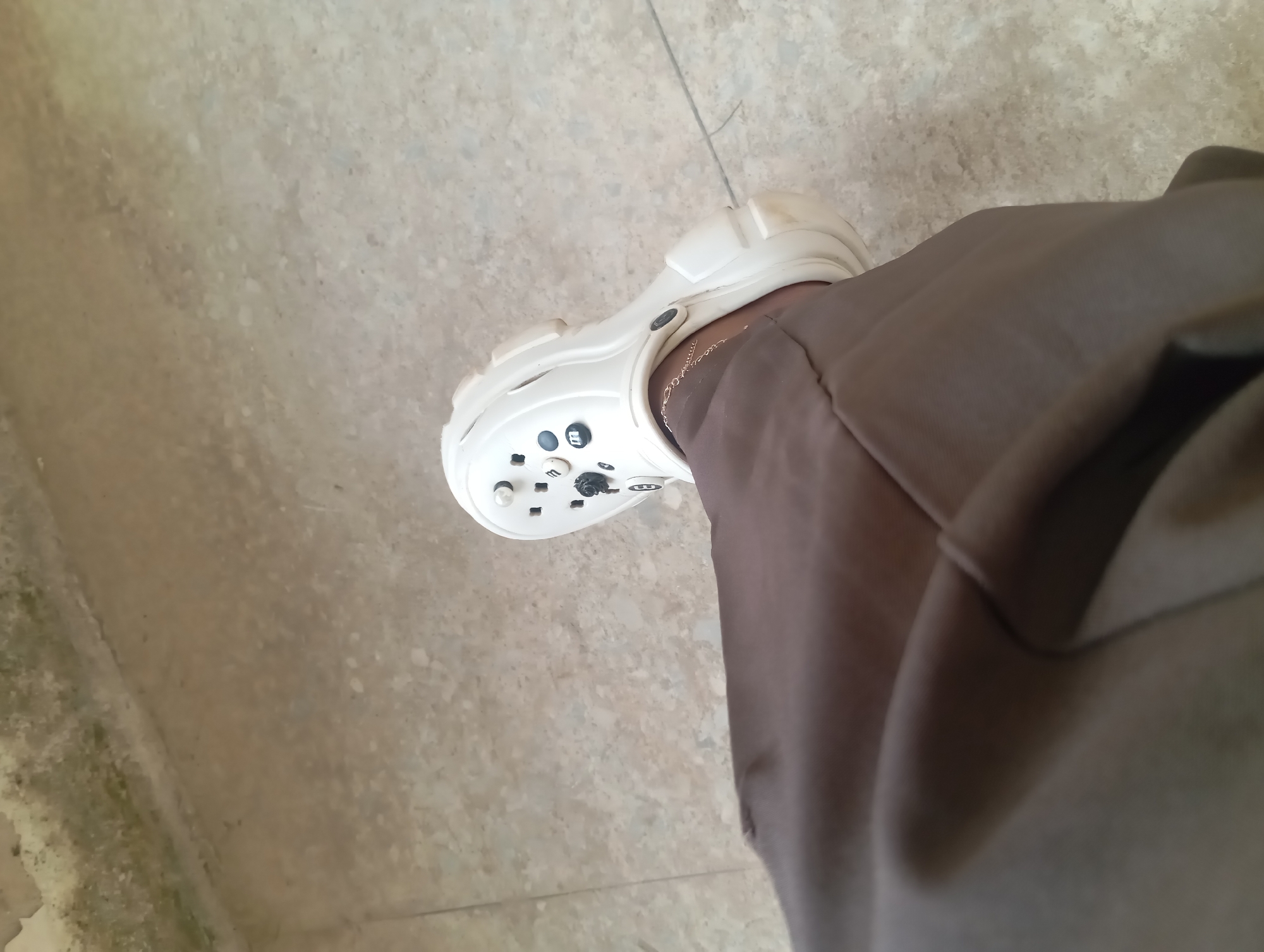 A person's left leg and foot, clad in brown pants and a white platform Croc, are visible from a high angle. The Croc features black decorative buttons and a black strap. A delicate gold anklet is worn on the ankle. The setting appears to be outdoors, on a concrete or stone surface with patches of discoloration and what looks like a thin wire or crack running across it. A light-colored, textured wall or curb is visible in the upper left corner of the frame, exhibiting signs of age and weathering. The lighting is somewhat diffused, casting subtle shadows.