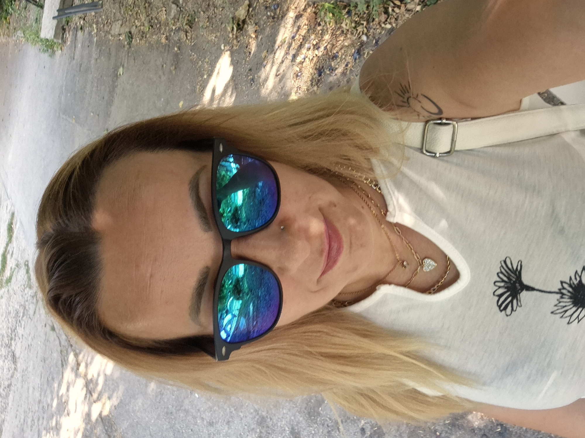A woman with blonde hair and sunglasses is standing outdoors. She is wearing a white shirt with a floral print and a necklace. She has a tattoo on her arm. The background is a pathway with trees and leaves. The lighting suggests it is daytime.