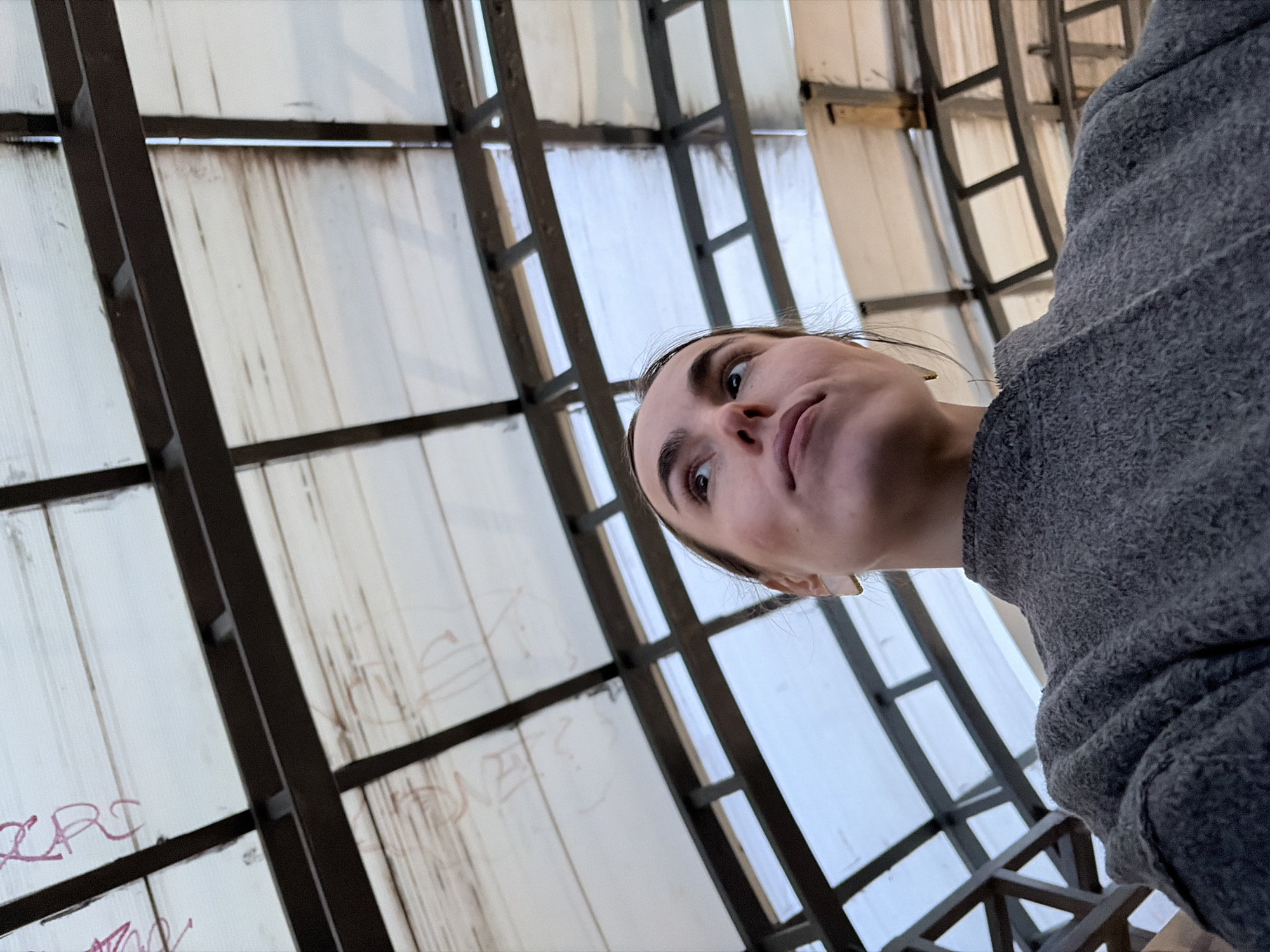 A low-angle, tilted shot depicts a person looking up at a ceiling structure. The ceiling appears to be constructed of translucent panels with a dark metal grid-like framework. Some panels have what appears to be writing or graffiti on them. The person is wearing a gray fleece or sweater. The location is Izhevsk, Russia.