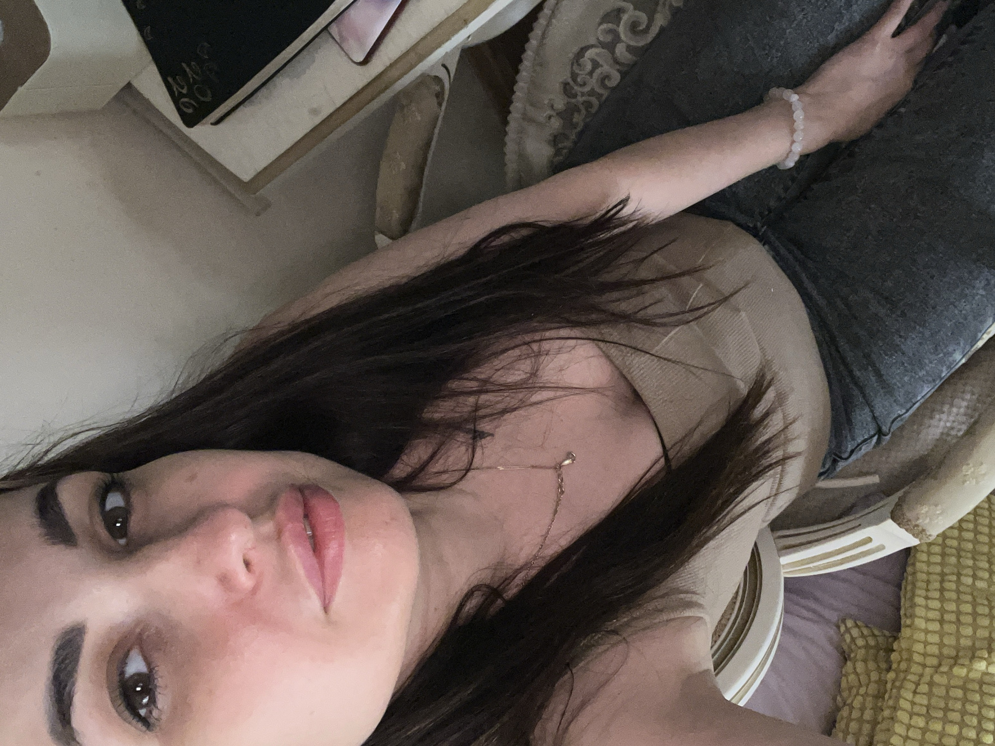 A young woman with long, dark hair is captured in a close-up, high-angle selfie. She is wearing a beige tank top and grey jeans. Around her neck is a delicate gold chain with a small pendant. On her left wrist, she wears a bracelet made of light pink beads. In the background, a desk is visible with a black notebook containing handwritten text. The environment appears to be indoors, possibly a bedroom or living area, with soft lighting.