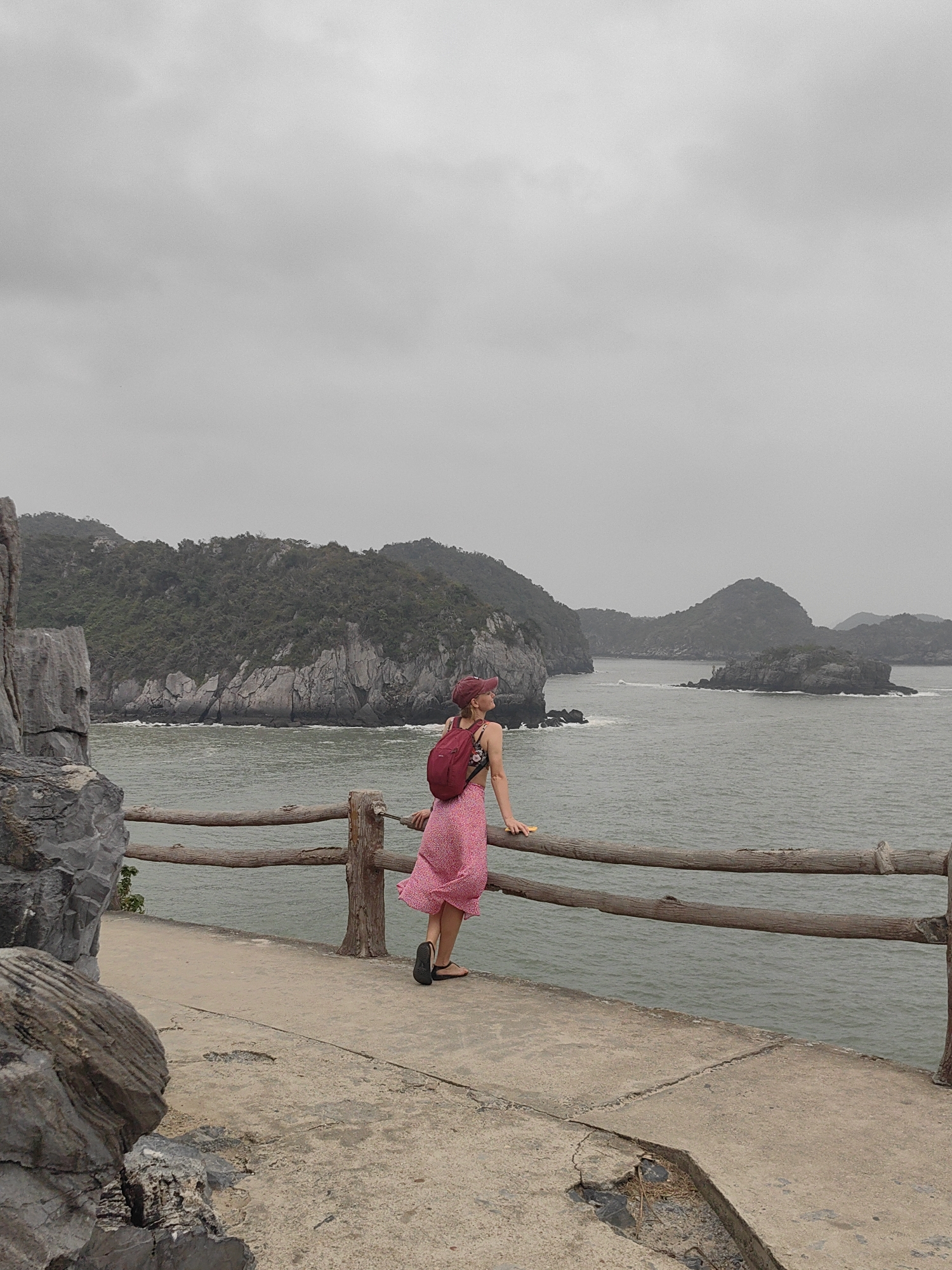 A medium shot captures a woman looking out at an overcast seascape. The woman, wearing a pink floral dress, a red cap, and a red backpack, leans on a weathered wooden railing. She is situated on a concrete walkway bordered by rocky formations on one side and the sea on the other. The sea is calm, and several islands rise from the water in the background, shrouded in mist. The sky is uniformly grey. The scene is located in the city of Hải Phòng, Vietnam.