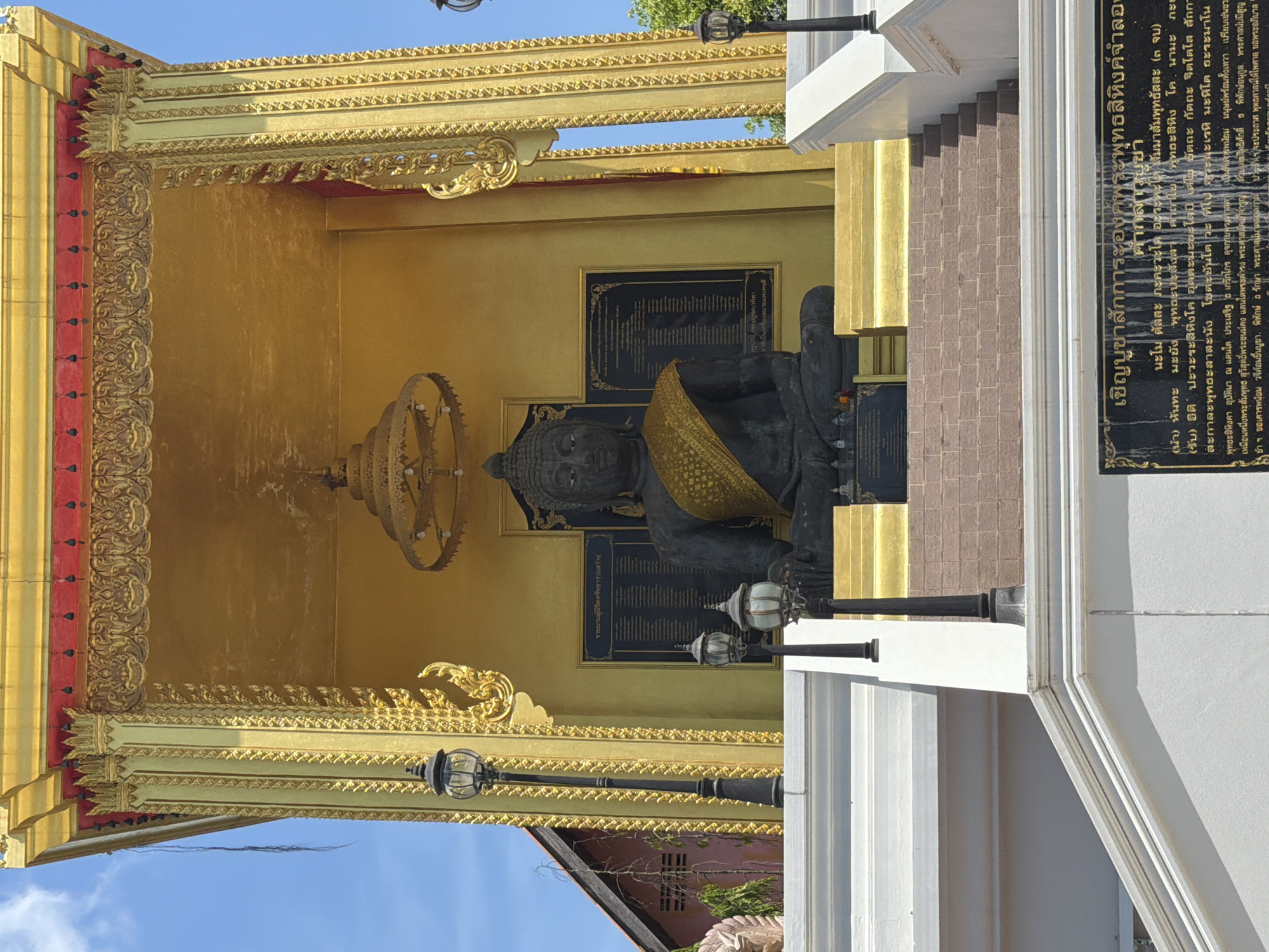 This image captures a serene outdoor scene at the Wat Phra That Mae Nam Temple in Baan Mae Nam, Thailand, under a bright, clear sky. The central focus is a large, dark stone Buddha statue seated within a golden, ornate shrine. The Buddha is depicted with a calm expression, adorned with a golden sash draped over its left shoulder. The shrine itself is richly decorated with intricate gold carvings and moldings, featuring a golden canopy above.
In front of the shrine, a series of white-tiered steps lead up to the statue, interspersed with brown brickwork on the lower levels. Several black lampposts with lantern fixtures are positioned on the steps and around the base of the shrine, suggesting the area is illuminated at night. Behind the Buddha, a dark plaque with white text is mounted on the golden wall, likely containing religious inscriptions or historical information.
To the left of the shrine, a portion of a traditional Thai building with a dark roof and reddish-brown walls is visible, with a leafless tree branch extending towards the shrine. In the foreground, more white architectural elements, possibly part of the temple's exterior or an entrance, are visible, including a large black plaque with Thai script. The overall atmosphere is one of tranquility and reverence, characteristic of a Buddhist temple.