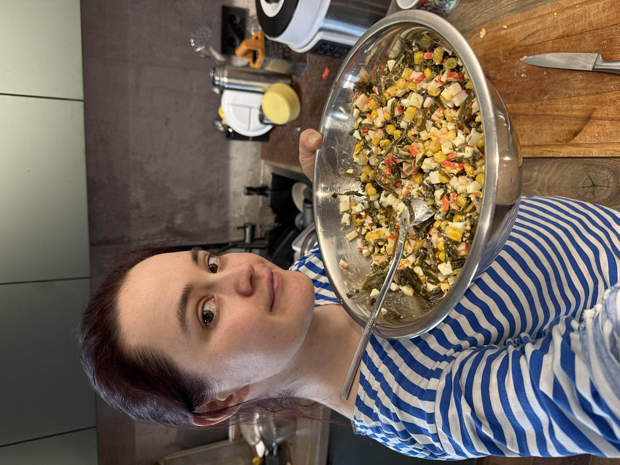 A person is holding a stainless steel bowl filled with a salad consisting of seaweed, corn, eggs, and imitation crab meat. A spoon is visible in the bowl. The person is wearing a blue and white striped shirt. A cutting board with a knife on it is to the right of the bowl. In the background, there is a kitchen counter with various appliances. The scene appears to be indoors. The location of Izhevsk, Russia is not discernible from the image.